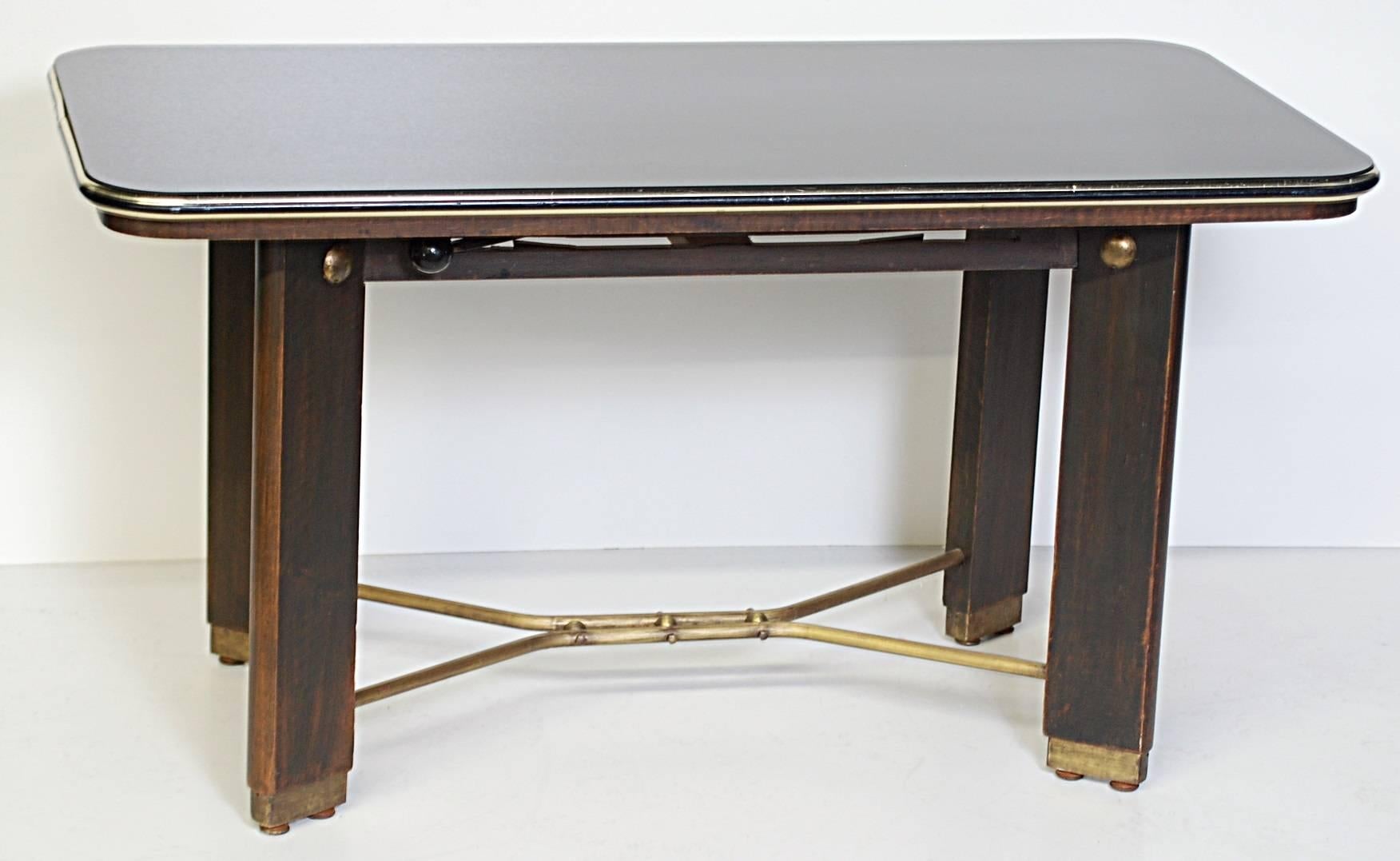 Unusual coffee table with a dark wood base, brass stretchers and brass end caps. Blackened glass top with brass trim. The mechanism is actuated by the lever raises the height of the top of the table. 28.5 inches extended - regular measurements below.