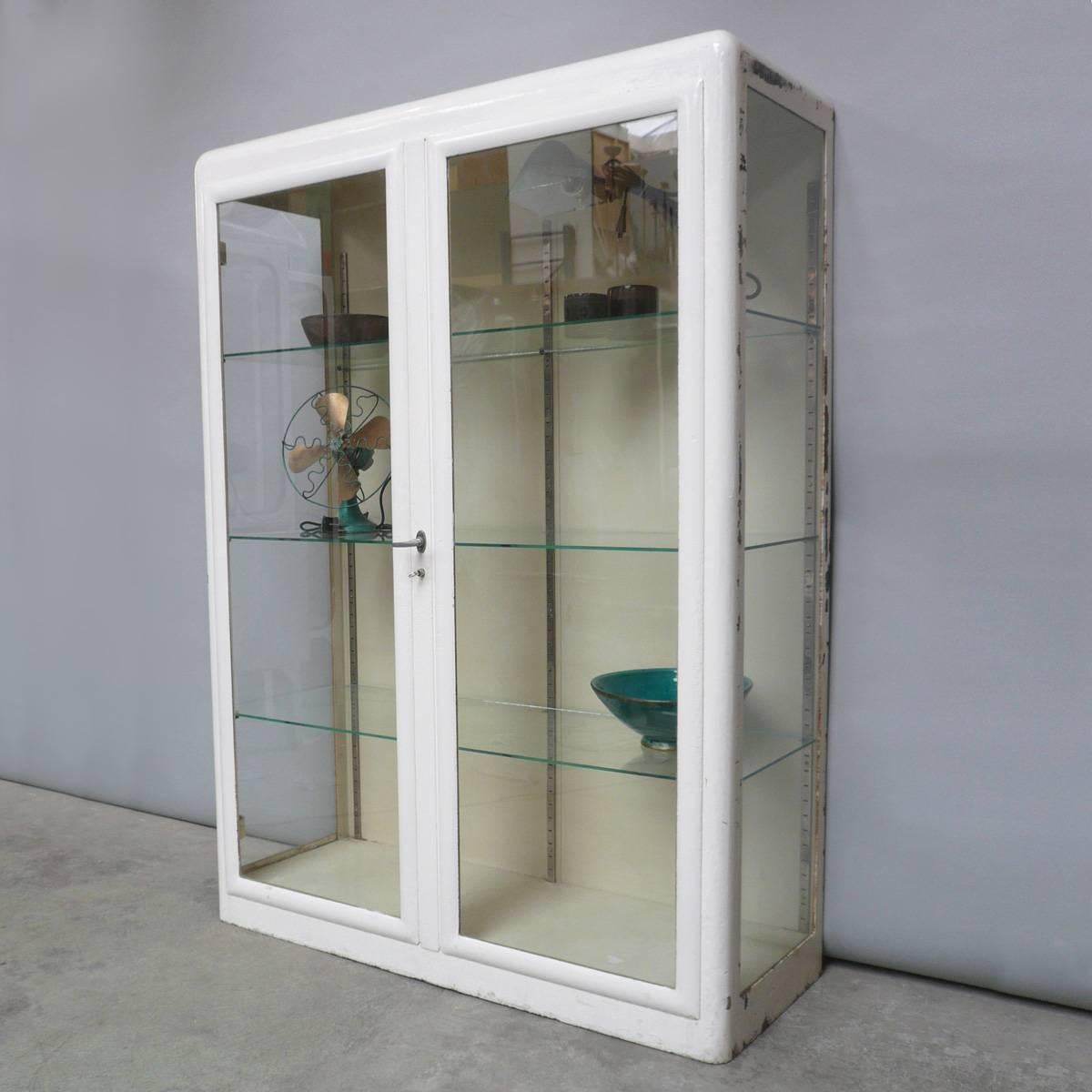 This vintage cabinet was produced in Hungary during the 1930s. The cabinet is made of steel with three glass shelves. The unit features two opening doors that lock with a key. The 150kg cabinet has a height of 170 cm and length of 123 cm. The item