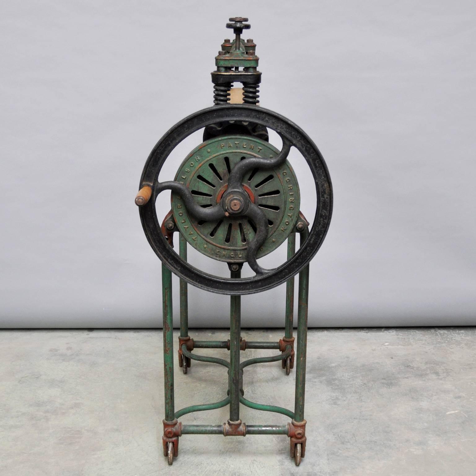 A large iron framed manual machine with two cylinders for rolling and pressing washed clothes, connected by cogs and manually operated by turning a side wheel with a large crank handle.
 