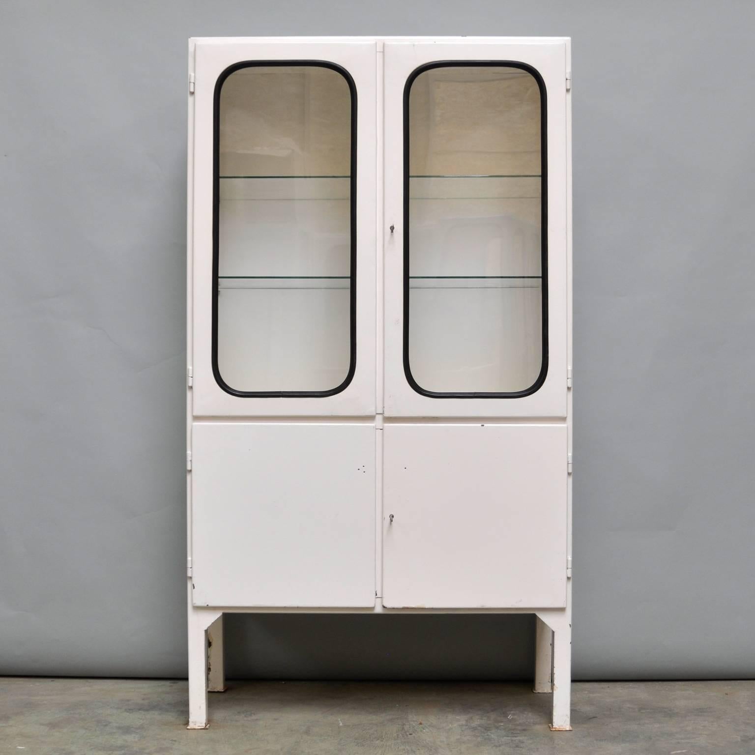 This medicine cabinet was designed in the 1970s and produced in 1975 in Hungary. It is made from iron and antique glass, with the glass panes held in place by a black rubber strip. The cabinet features two adjustable glass shelves and functioning
