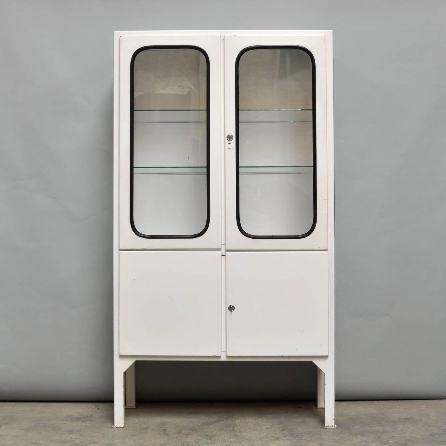 This medicine cabinet was designed in the 1970s and was produced circa 1975 in Hungary. The piece is made from iron and glass, and the glass is held by a black rubber strip. The cabinet features two adjustable glass shelves and a functioning lock.