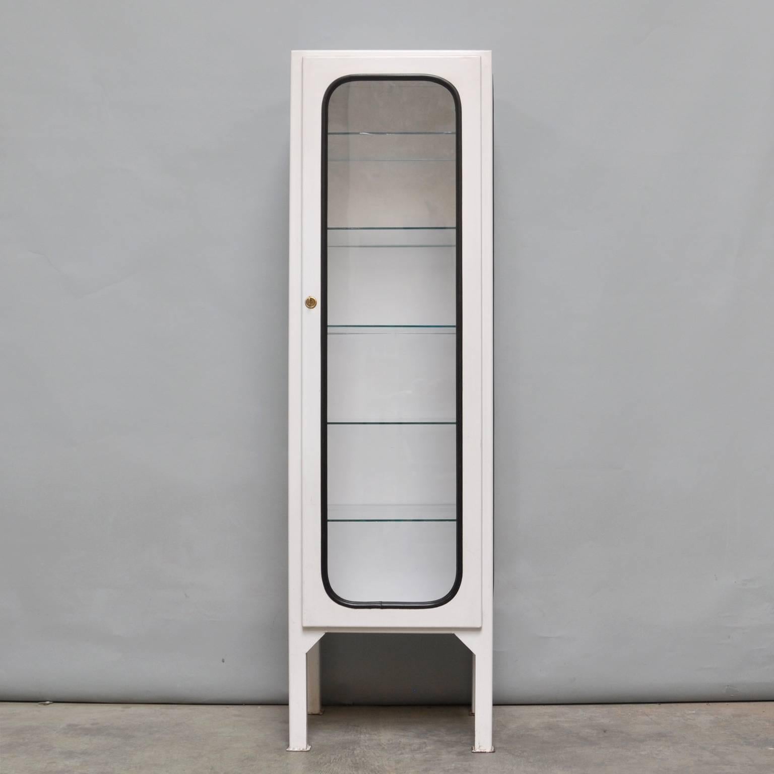 This medicine cabinet was designed in the 1970s and produced in 1975 in Hungary. It is made from steel and glass, with the glass panes held in place by a black rubber strip. The cabinet features five adjustable glass shelves and a functioning lock.