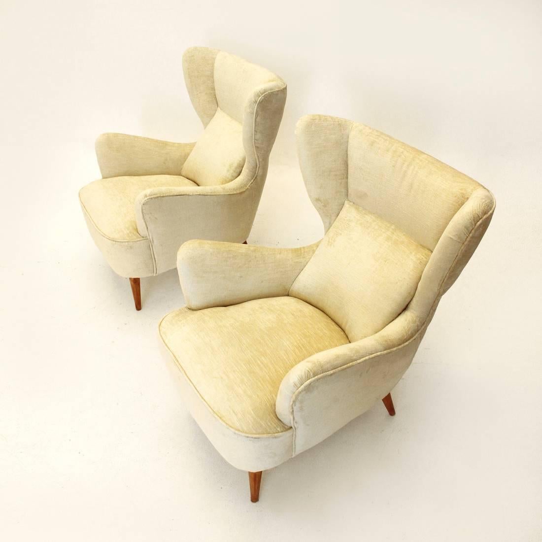 This pair of wingback armchairs was made in Italy in the 1950s. They feature wooden frames, conical legs and cream-colored velvet upholstery.