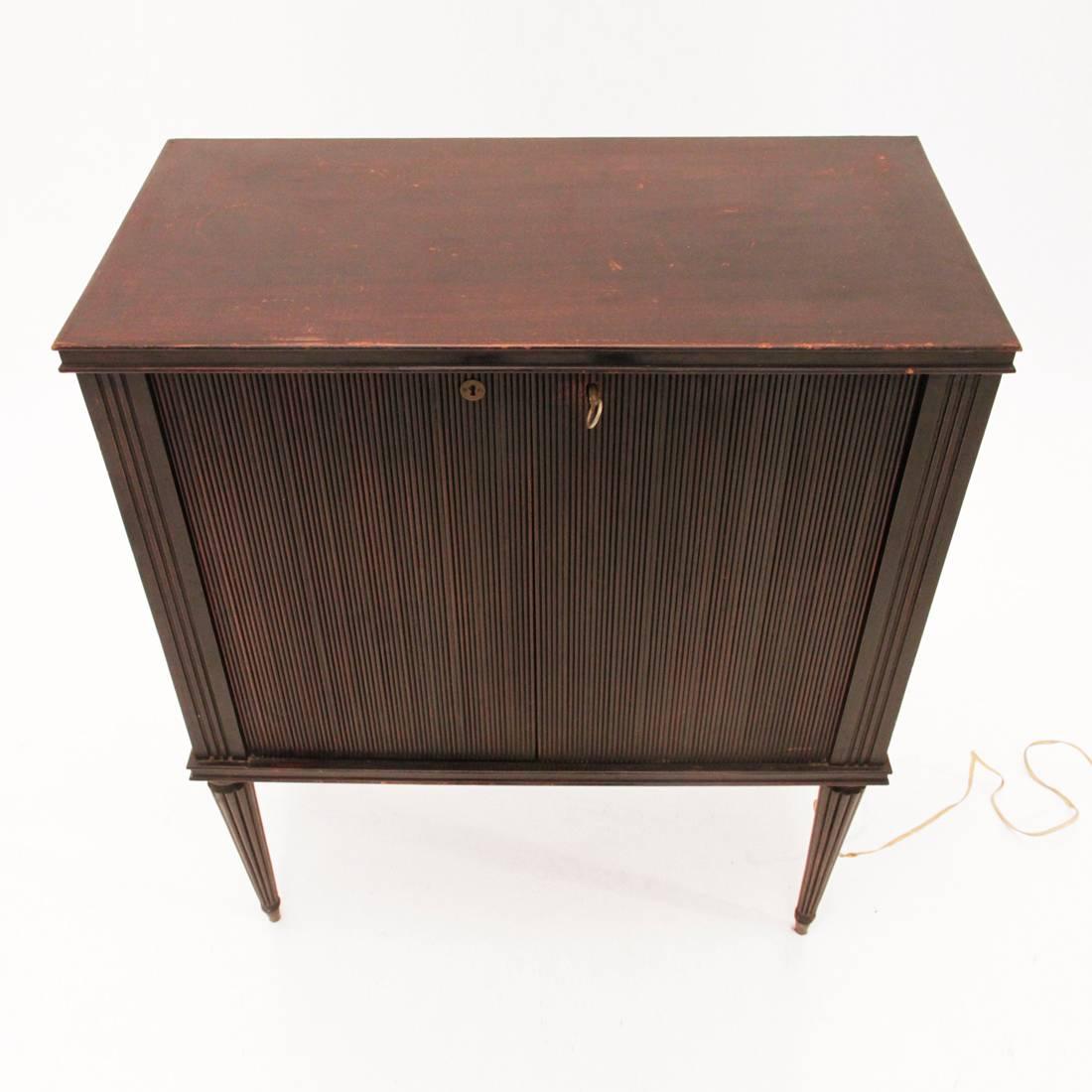 This wooden bar cabinet was manufactured in Italy during the 1940s and features two wooden ribbed doors, an interior magazine bar surrounded by mirrors, an interior electric bulb for lighting and tapered legs with brass feet.