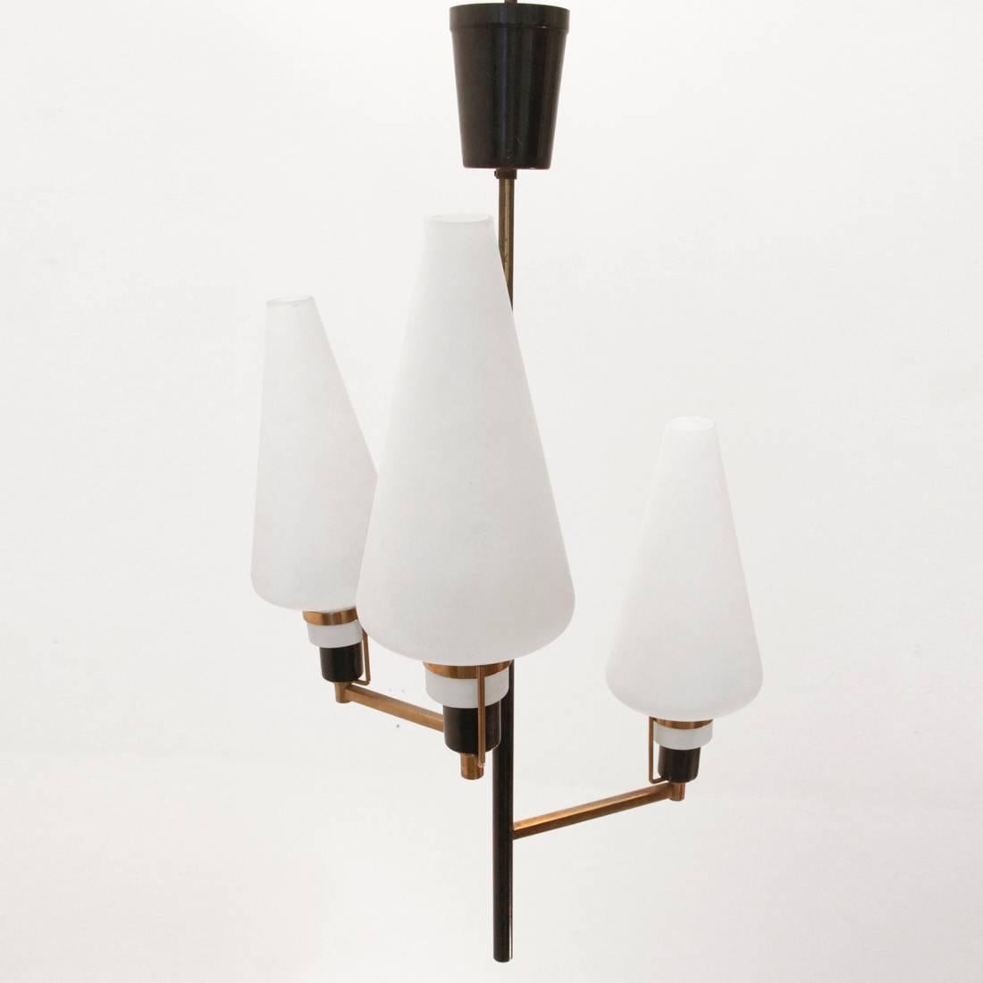 This chandelier was produced in Italy during the 1950s. It features a three-armed brass structure with black metal elements and white opal glass cone-shaped shades. Good vintage condition.