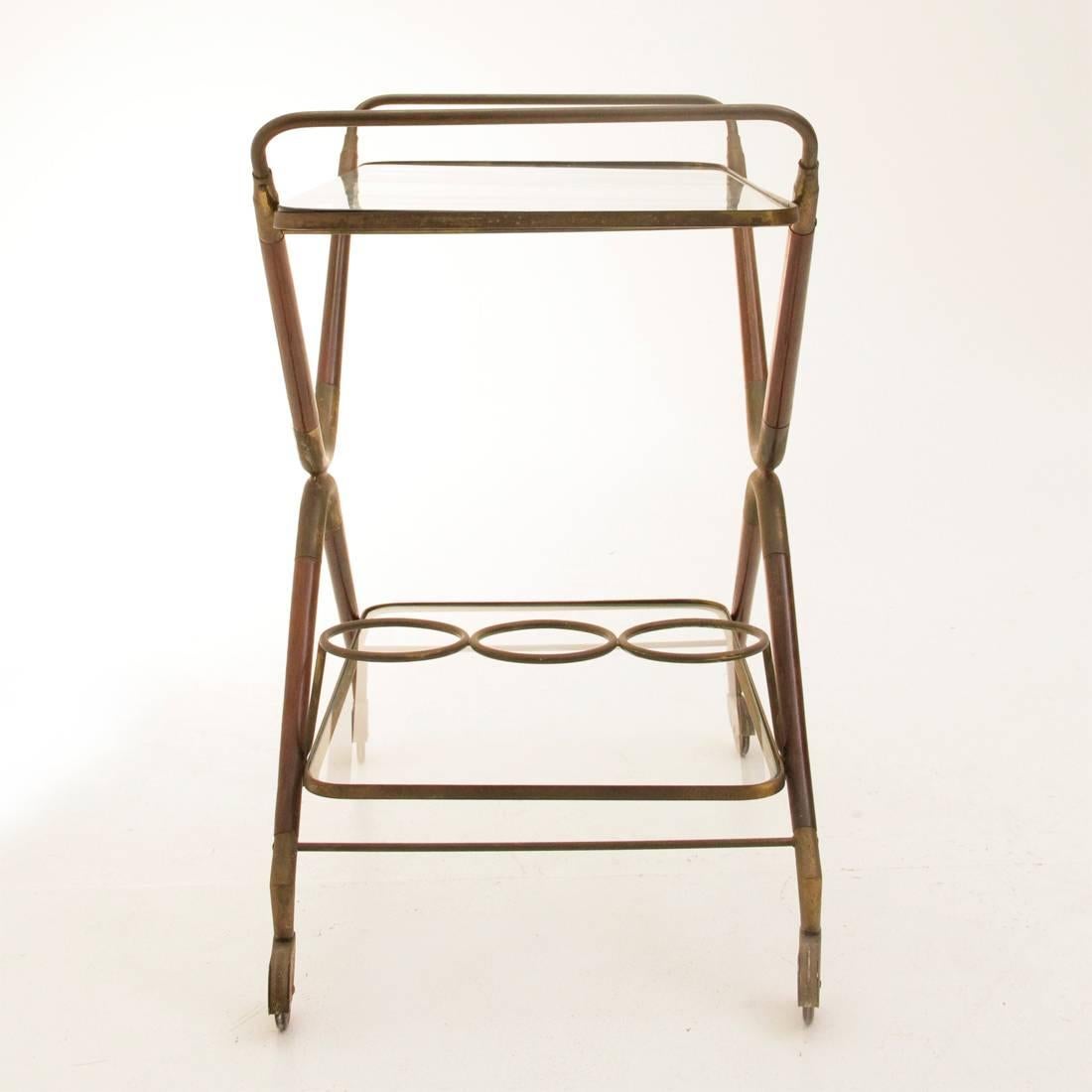 This lunch trolley designed by Cesare Lacca in the 1950s features a double C-structure in inverted brass and lacquered wood, supporting two shelves with brass edging, and a glass top, all sitting on brass and rubber wheels.