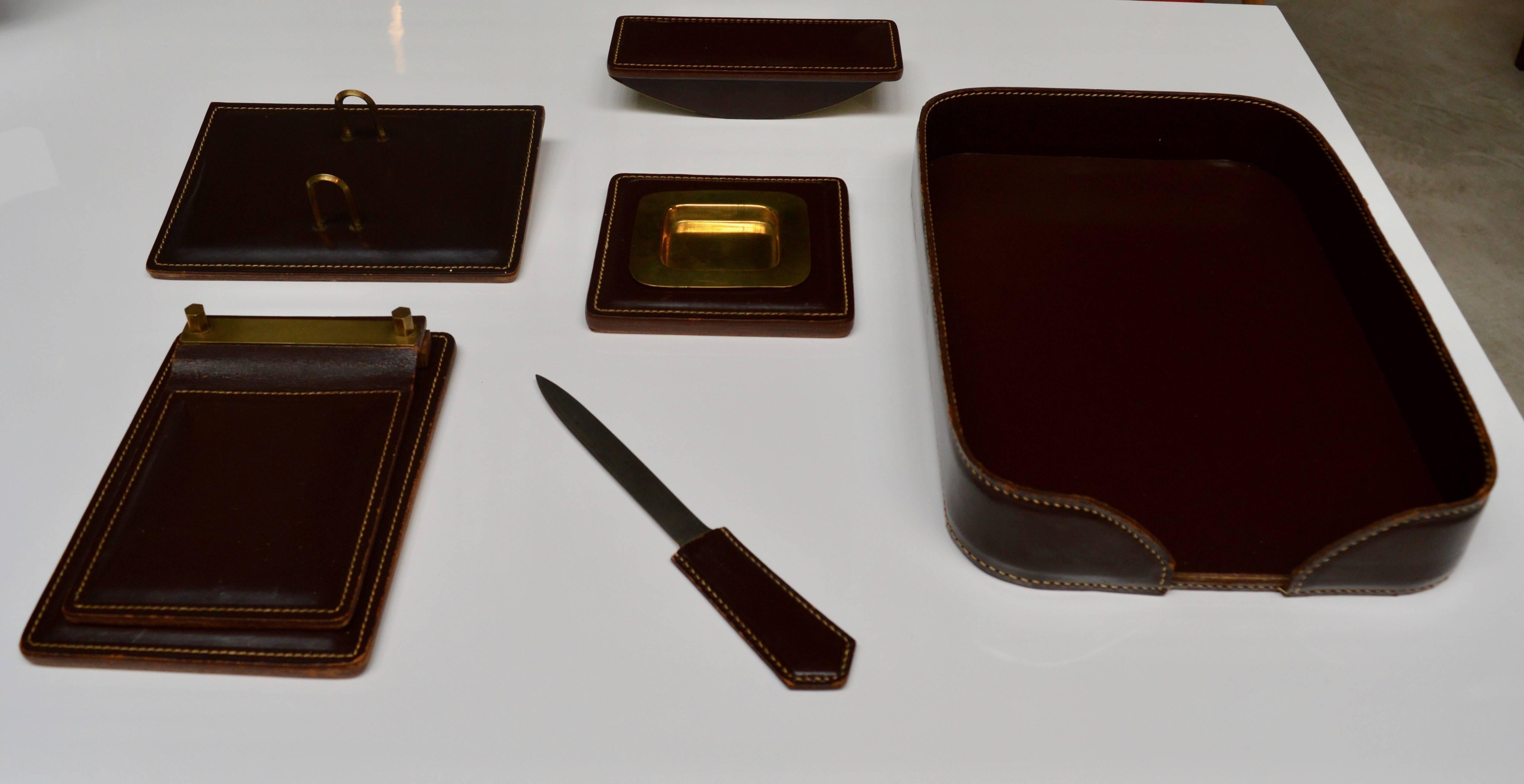 Handsome French leather desk set in the style of Jacques Adnet. Deep brown/burgundy leather with contrast stitching. Six-piece set includes letter tray, letter opener, ashtray/catchall with brass dish, calendar pad, notepad and ink blotter.