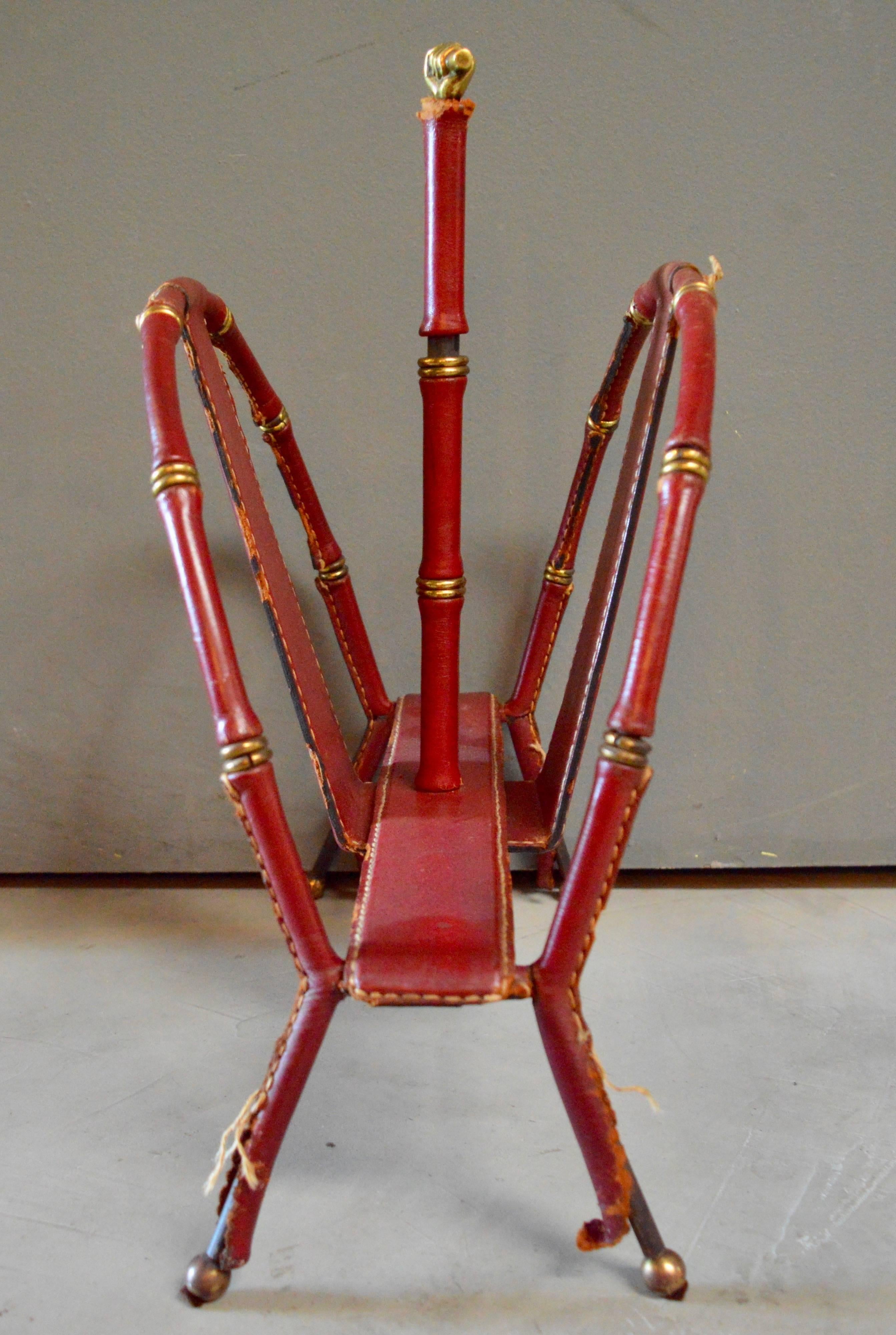Handsome magazine rack by French designer Jacques Adnet. Red leather wrapped around iron frame, with brass ball feet, brass trim and brass clenched fist. Signature Adnet contrast stitching. Original condition. Unrestored. Great collectors piece of