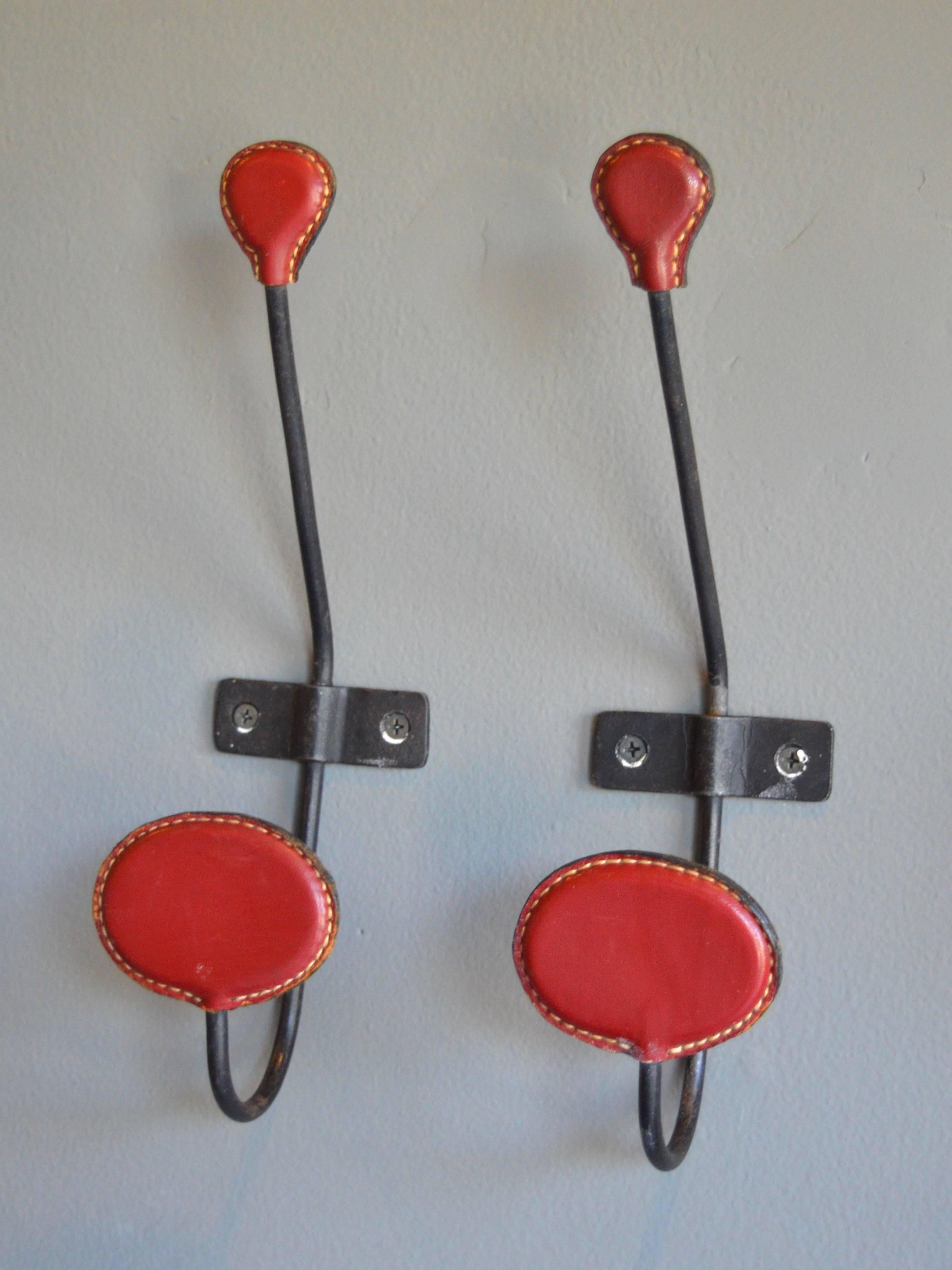 Handsome pair of coat hooks in the style of Jacques Adnet. Iron frame with deep red leather and stitching. The top of the hook has a small tear drop shaped arm and the bottom of the hook has a larger, ovular shaped arm. Good vintage condition. Sold