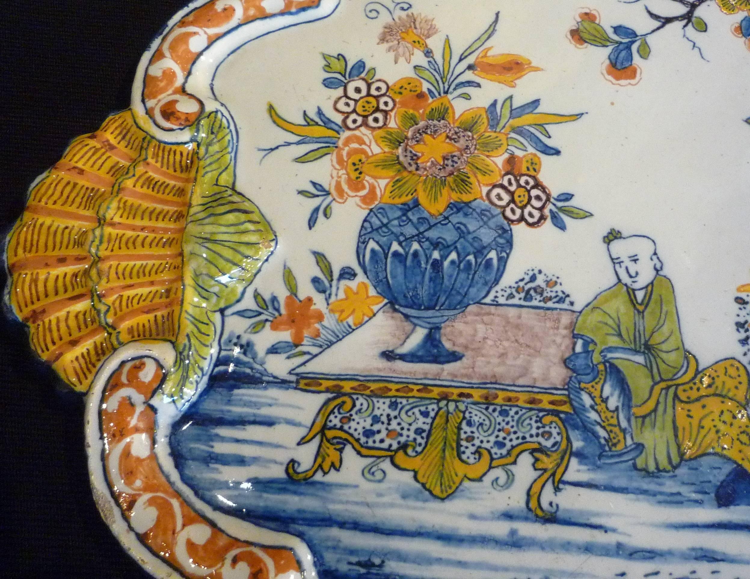 A large delft polychrome plaque with border of shells, the center with Chinese figures in a landscape, with birds, trees and flowers, first half of the 18th century. No restoration.