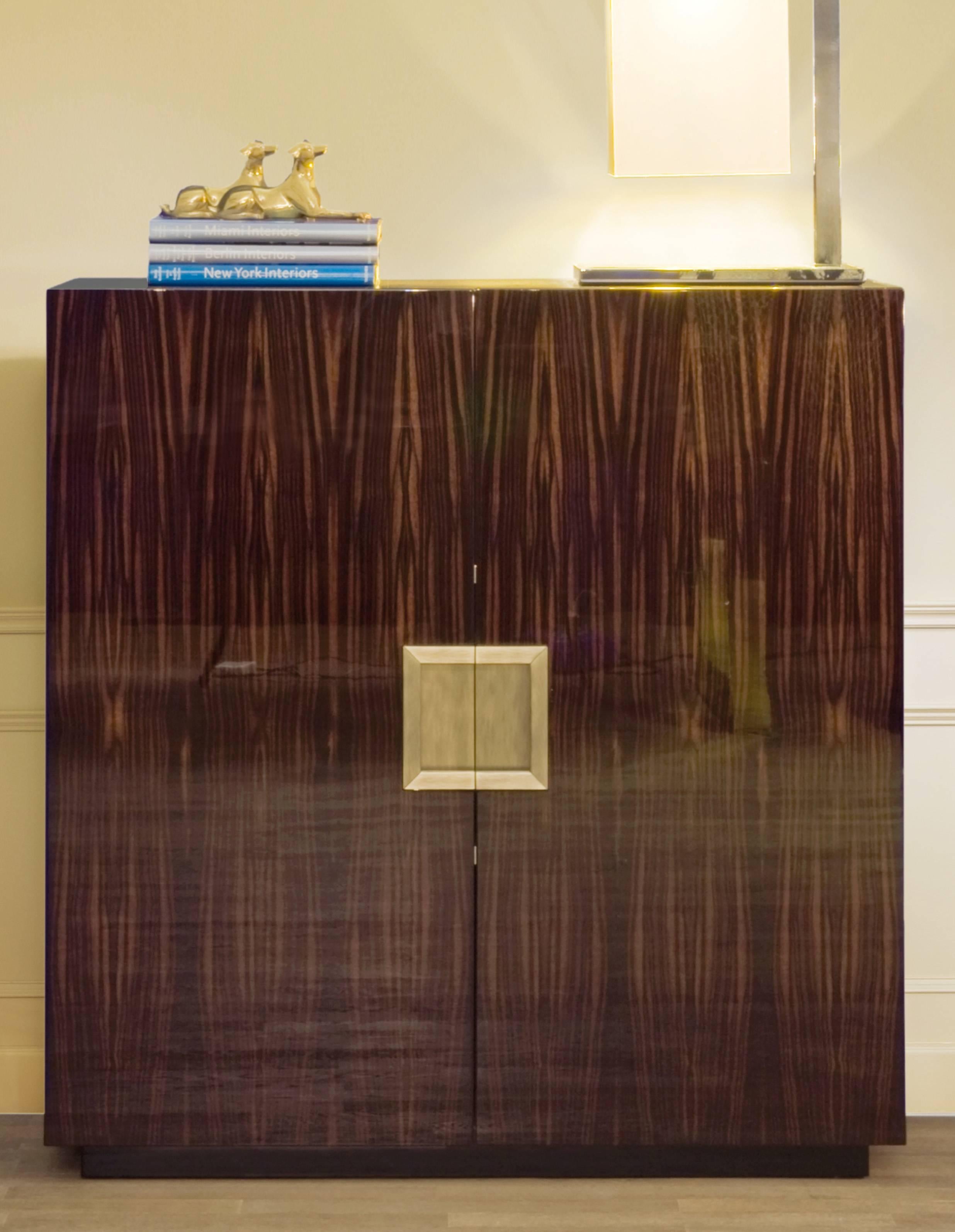 Cabinet with two doors, two internal shelves and metal handles, in the finishes gloss brass-plated, gloss chromed or bronzed. Macassar ebony veneered, in mat, semi-gloss or gloss finish with mat, semi-gloss or gloss maple interiors and gloss black