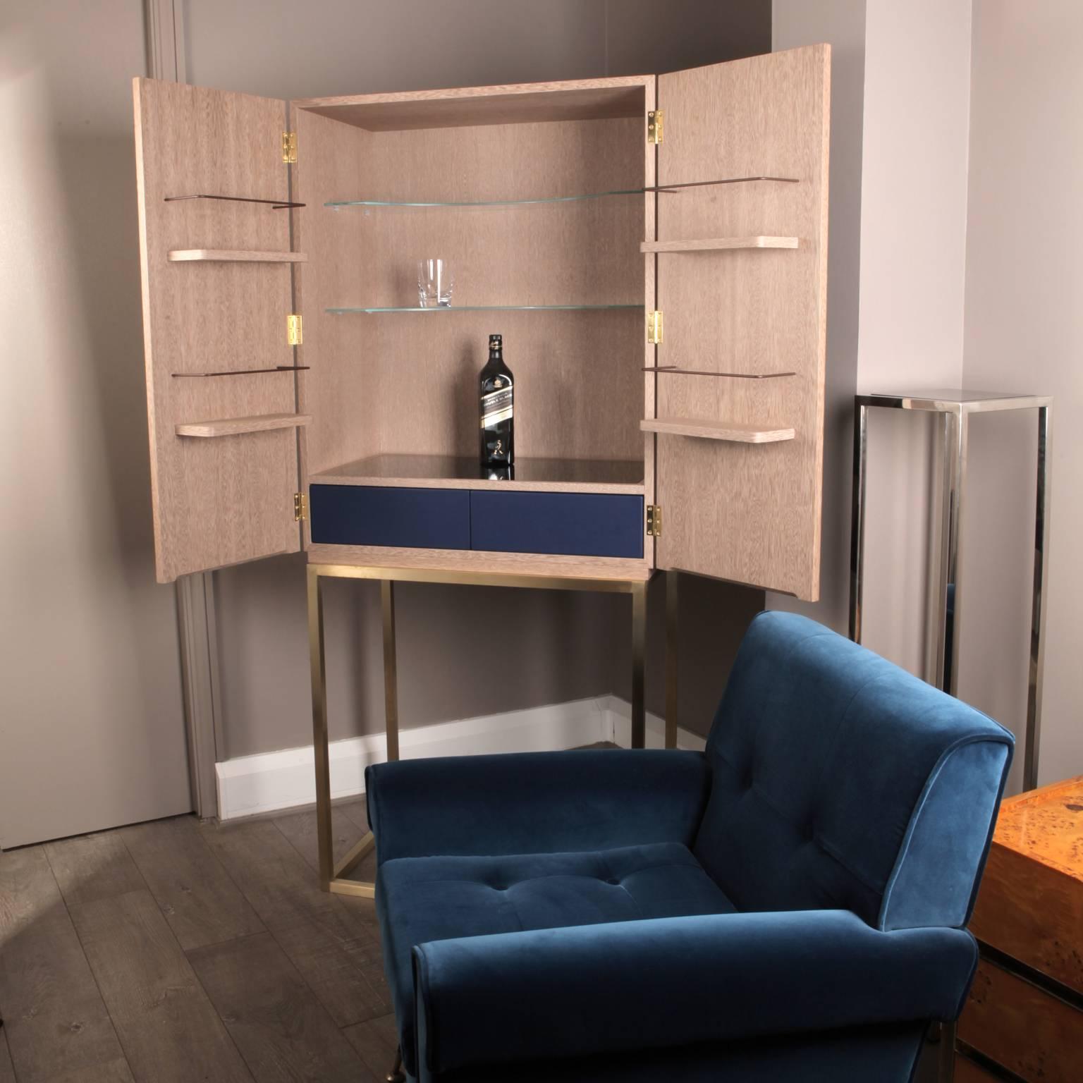Cerused wood carcase with brushed brass and bronze patinated brass triangle doors. the cabinet sits on a brushed brass frame. Two internal drawers upholstered in blue leather, glass shelves and bottle storage shelves.