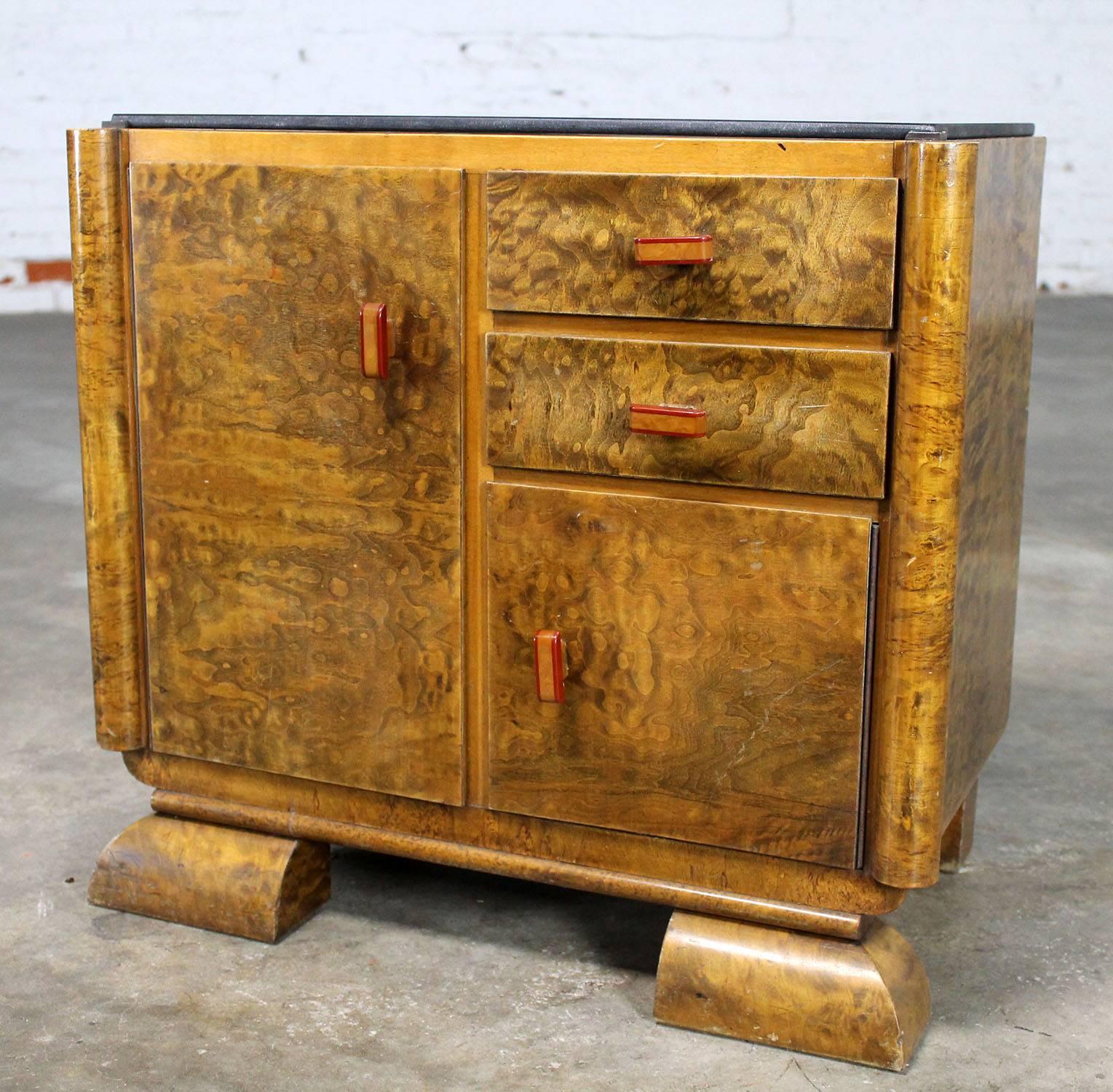 Wonderful petit sized Art Deco side cabinet or commode in gorgeous burl wood topped with a piece of black glass and accented with beautiful Bakelite handles. It is in fabulous vintage condition.

This is such a fabulous little Art Deco cabinet!
