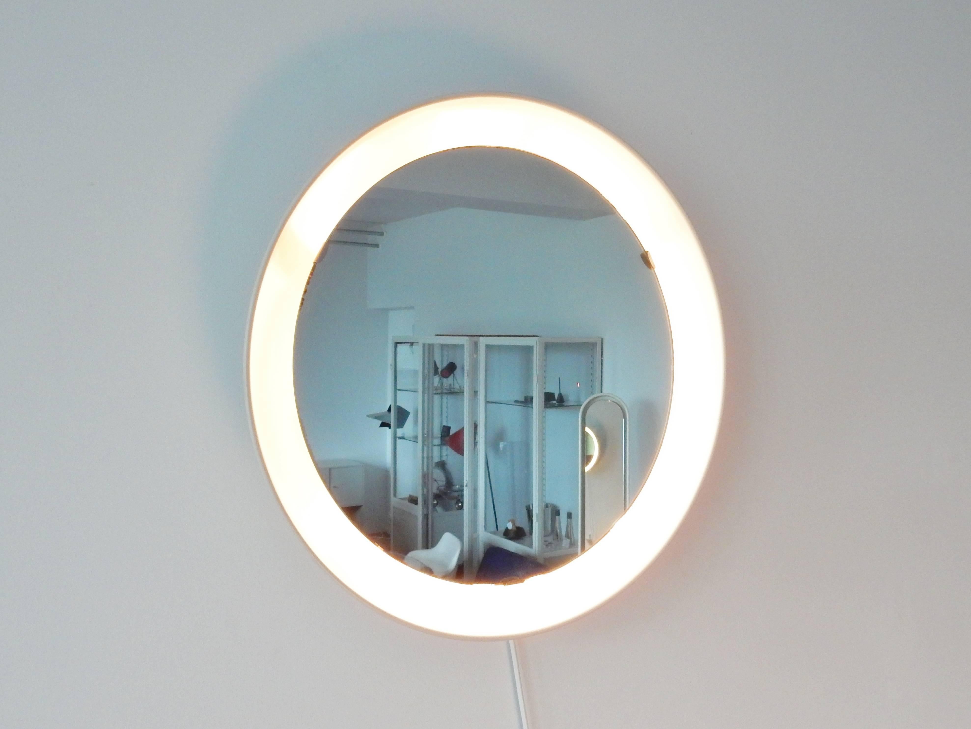 This mirror is quite a rare find. The short production period makes it a scarce product. The round glass mirror is placed into a large white lacquered aluminum round bowl that holds three-light bulbs. The light comes from behind the mirror and gives