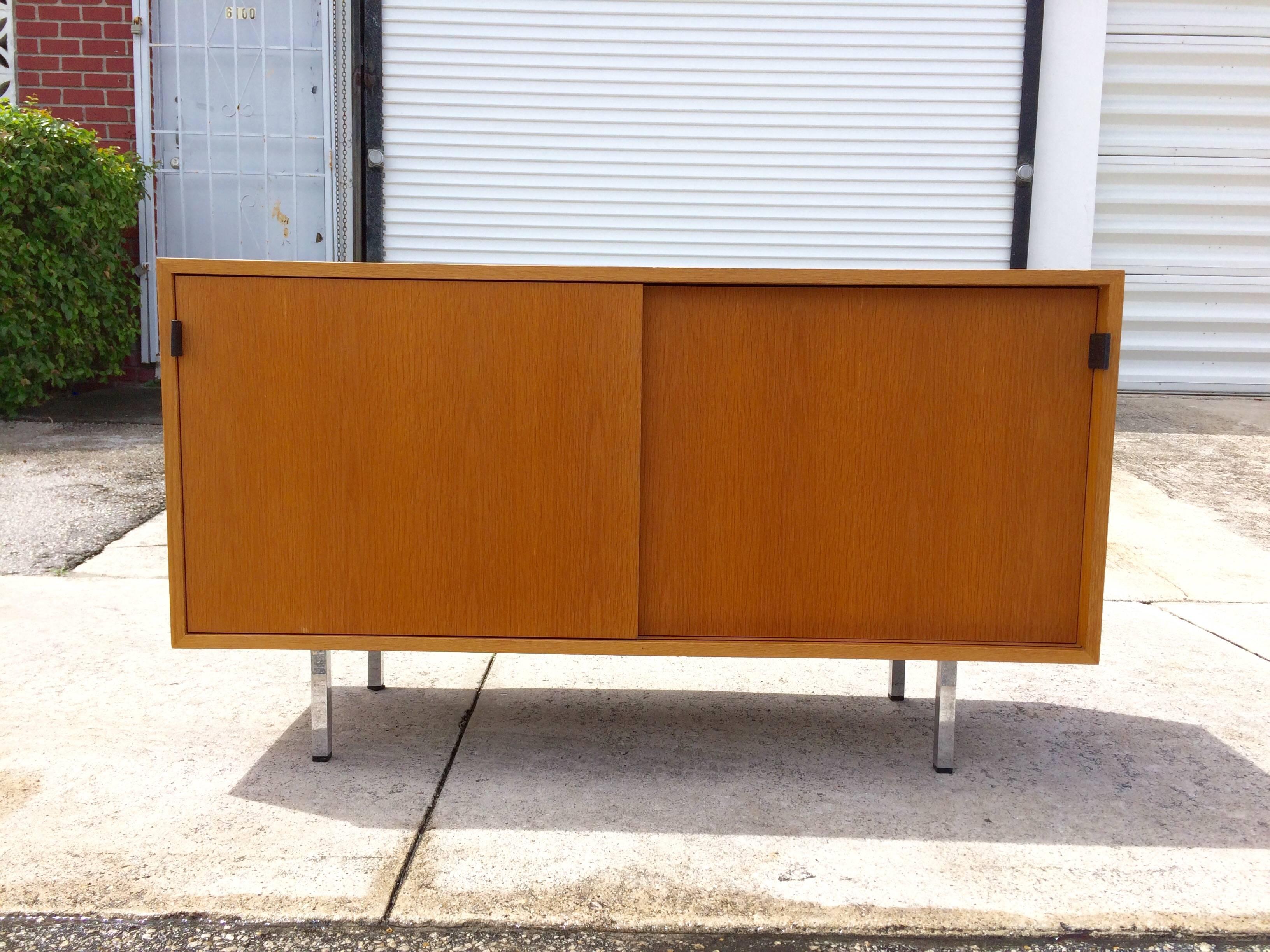 Credenza on four chrome legs, two sliding doors with black leather pulls. Behind the sliding doors are four shelves.