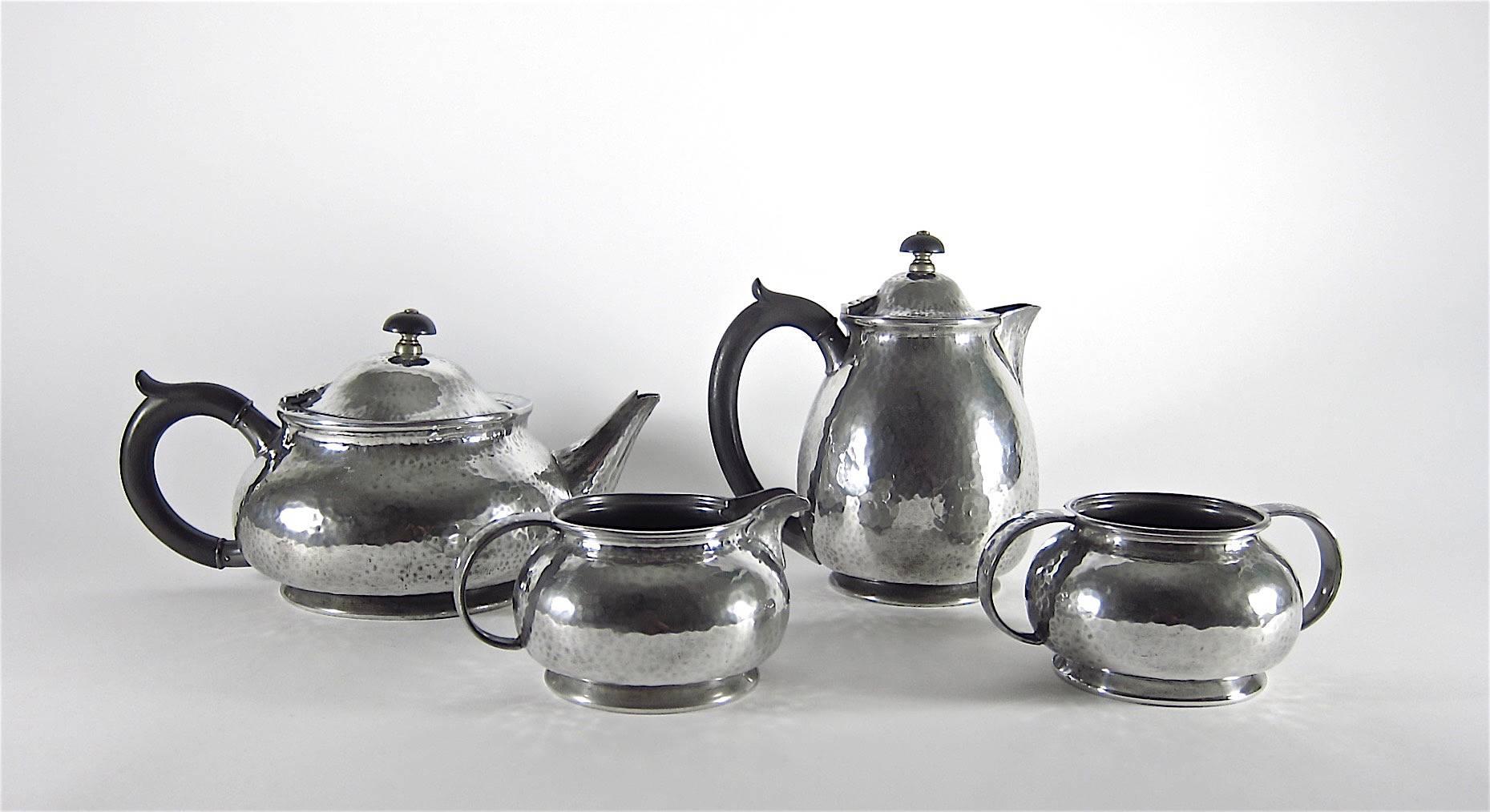 An early 20th century English Arts and Crafts pewter tea set comprised of four pieces: A teapot, hot water pot, cream pitcher and open sugar bowl. The larger pieces feature hinged lids with carved and turned handles and finials of ebonized wood.