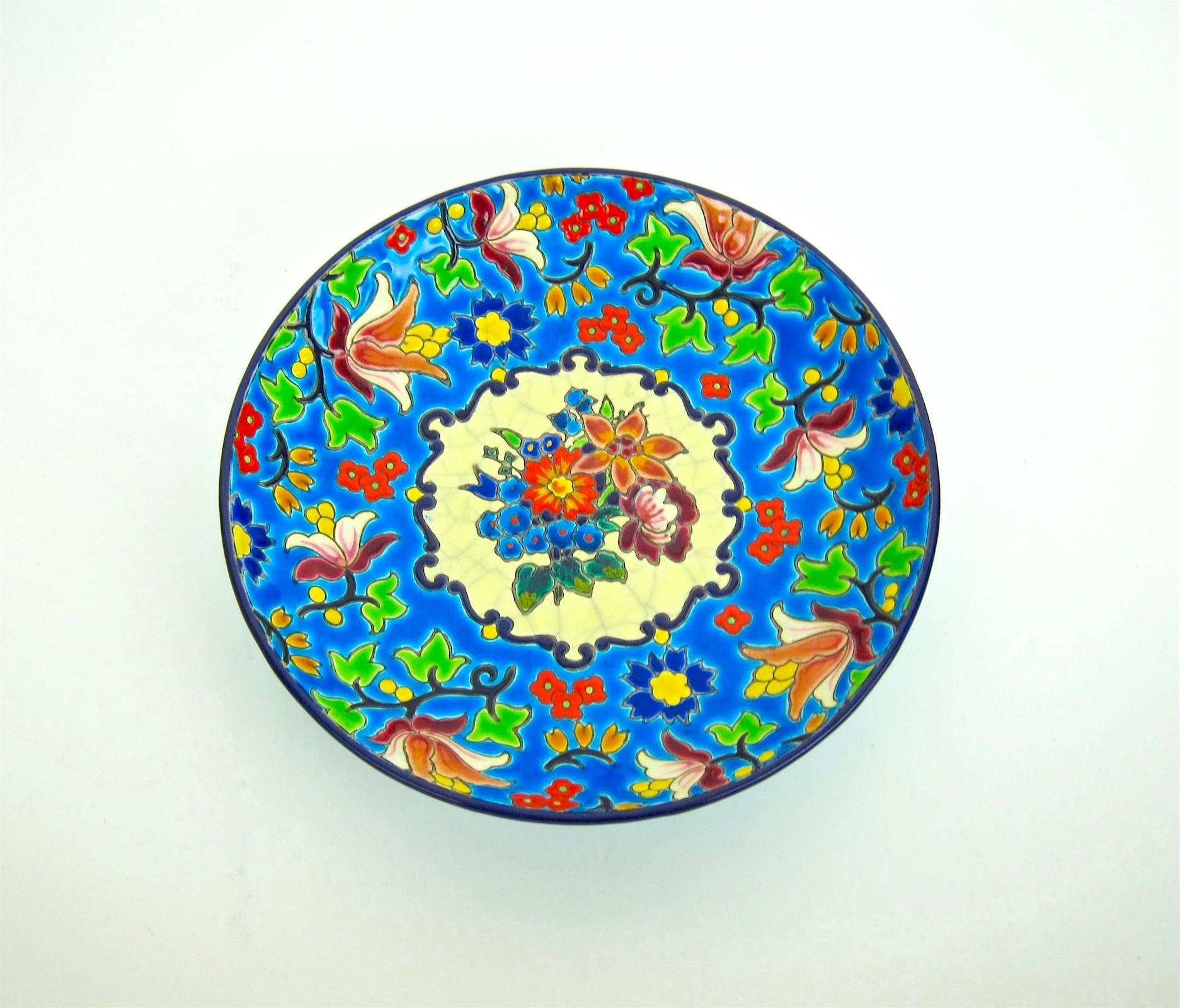 A hand-decorated French Emaux de Longwy pottery shallow bowl resting on a ring foot with an incised, cloisonné-style bright enameled glaze of multicolored blossoms with green leaves and black stems against a rich blue ground. The underside is glazed