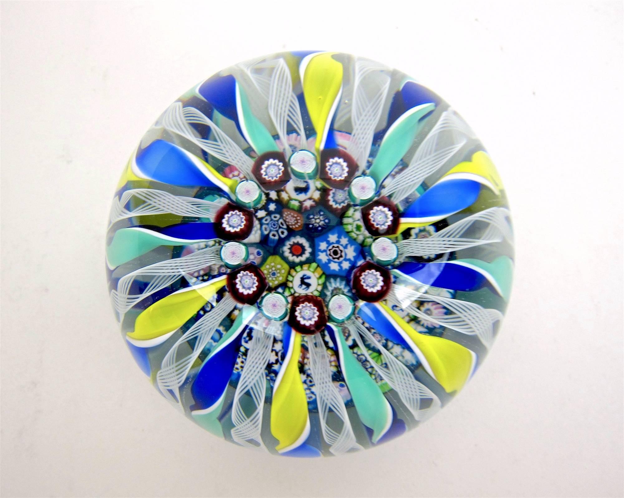 A large and colorful hand crafted studio glass paperweight from John Deacons of Scotland. The design is called a porthole with close packed complex millefiori and four silhouette canes interspersed with Clichy-style roses. Twisted ribbons of blues,