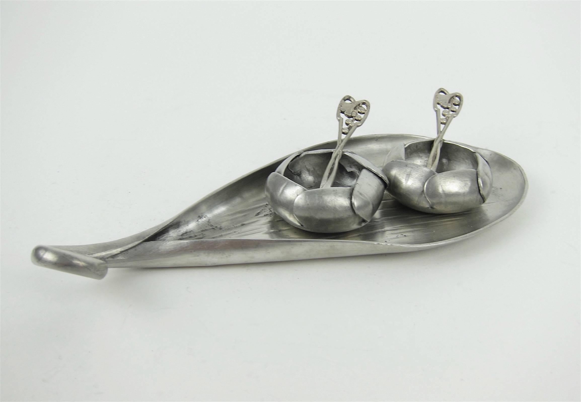 An exquisite early 20th century French Art Nouveau open salt and pepper set in hand-wrought and engraved pewter consisting of two floral-form salt cellars with overlapping petals, two spoons and an under plate with an elegantly swirled handle by
