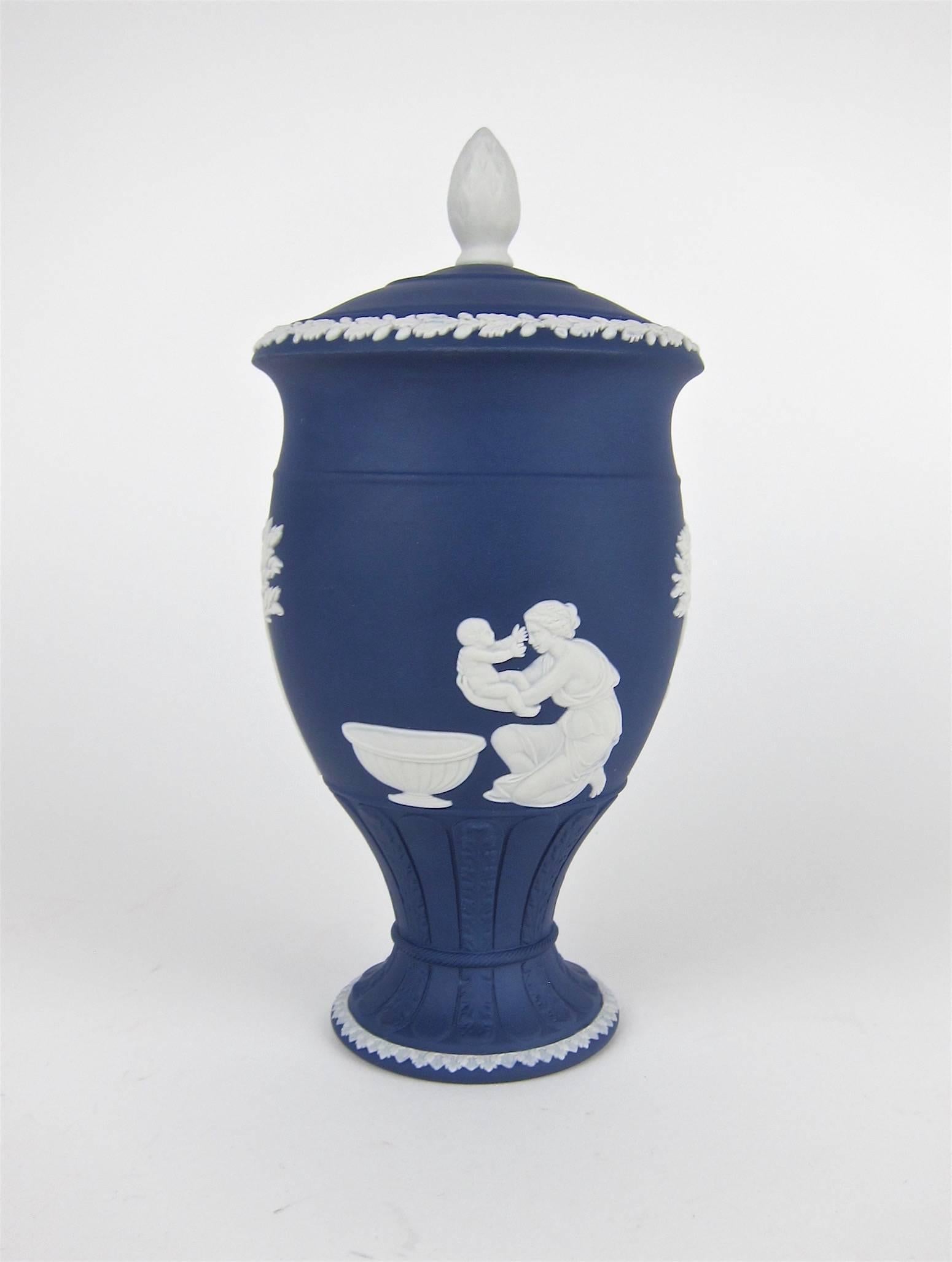 A covered vase in the shape of a Classical urn decorated with white on solid Portland Blue jasper ware from Wedgwood of England. The Neoclassical bas-relief decor depicts scenes of Achilles with his Mother interspersed with trees. The lid features a