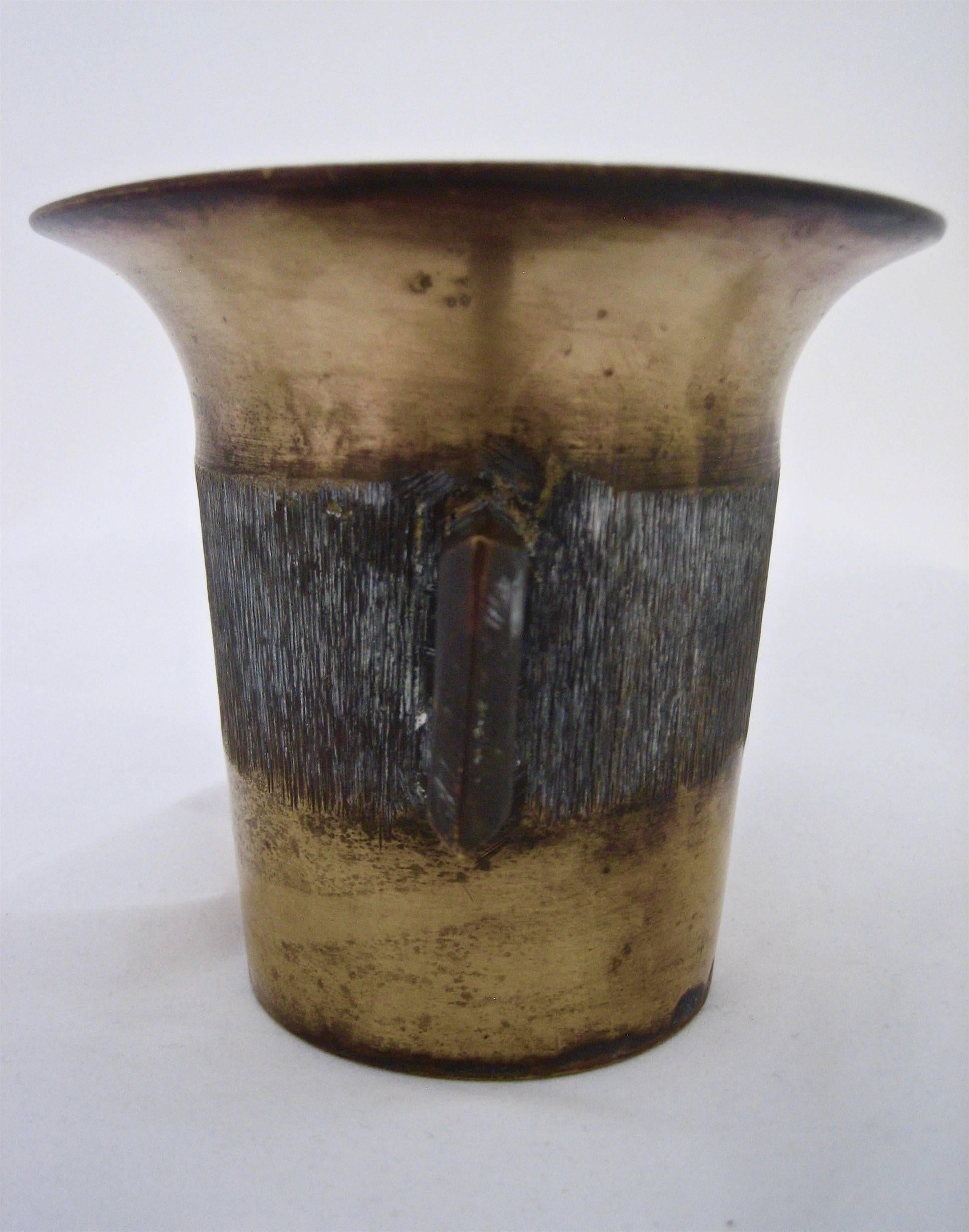 A 20th century small Trench art vessel with applied handles decorated with a scratched horizontal band. Substantial weight and a scored, circular groove pattern underfoot show the object was likely handcrafted from a spent shell casing. 

This