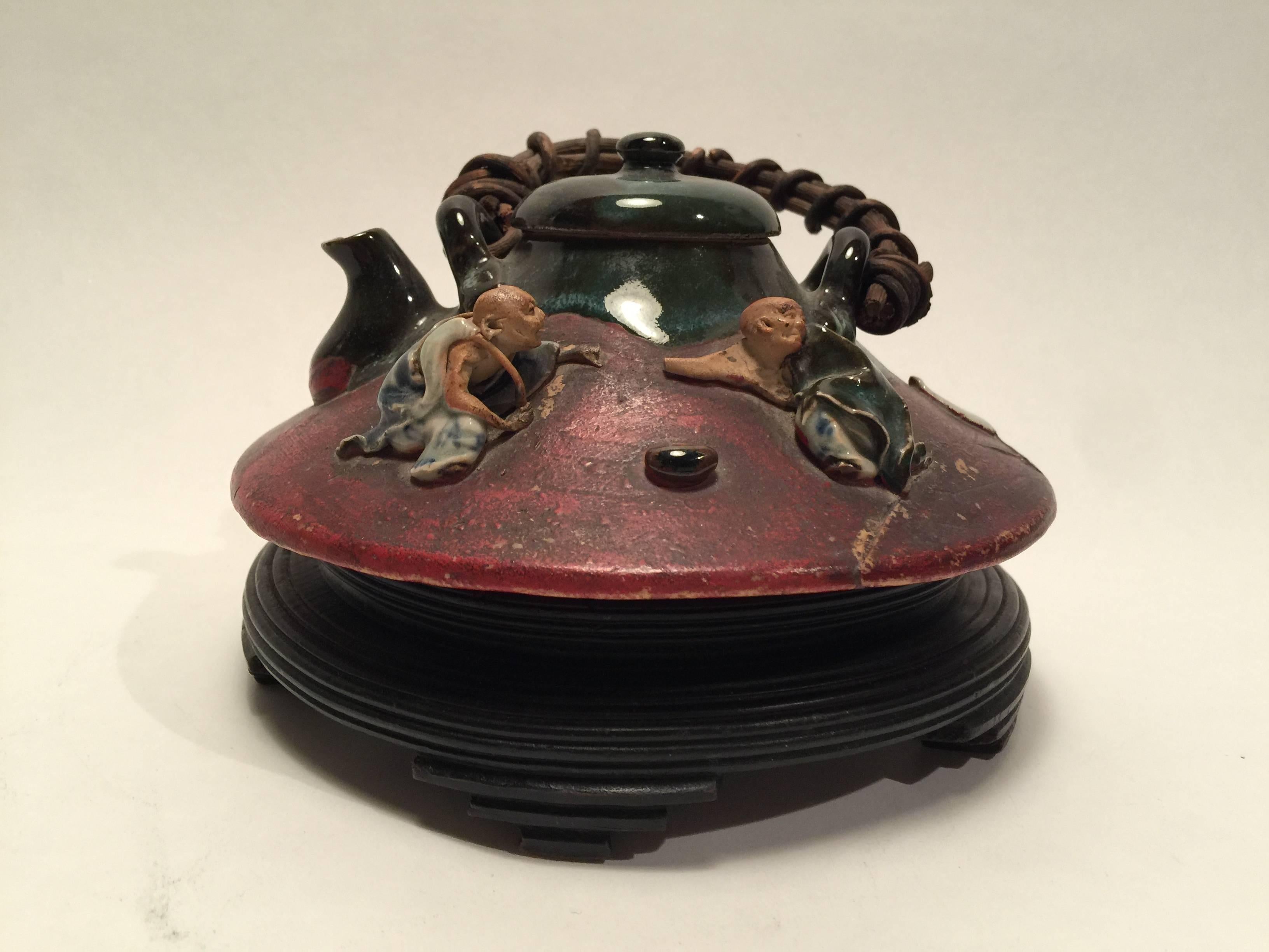 Two dimensional figures of men with expressive and detailed faces adorn the outer perimeter if this low tea pot.
There is a heavy glaze on the upper portion of the piece including the lid, while the lower portion is painted red. 
Several areas of