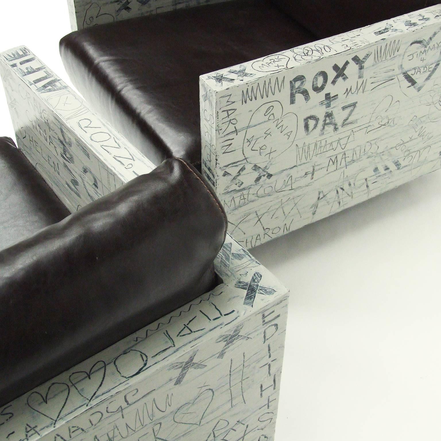 Pair of lounge chairs finished in a freeform hand-painted sweetheart graffiti design by Alan Fears.

Painted textured box frame upholstered in dark brown leather.

Ebonized plinth.

