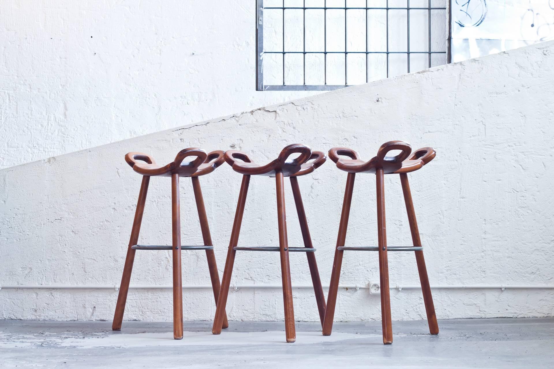 Brutalist bar stools in dark-brown stained beech. A curved T-shape with three handles. The curved form makes sure the stool has a stable seat, emphasized by the metal ring as footrest.