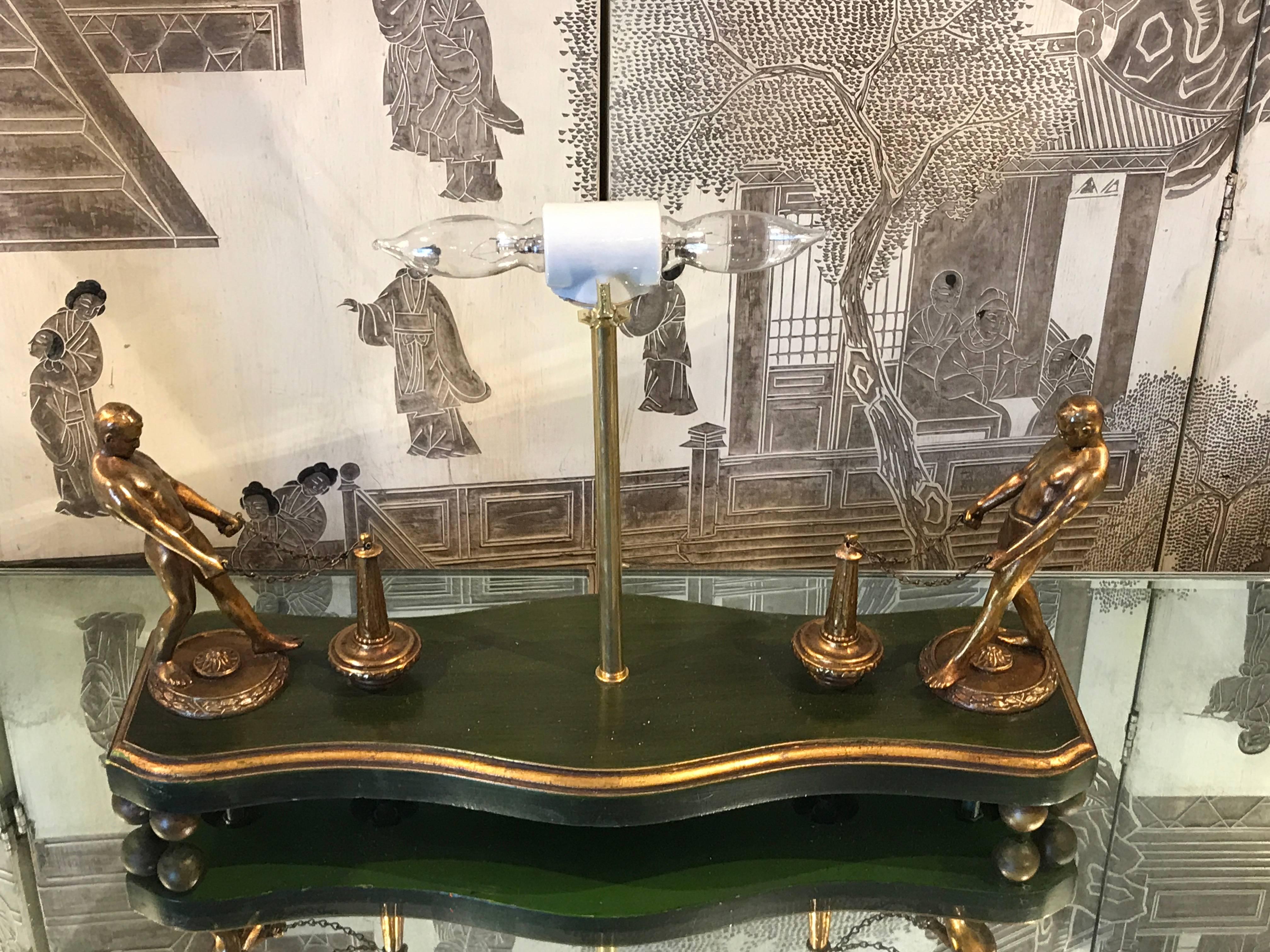 Art Deco Male figural lamp, with two draped male figures at each end chained to a post, raised on a green ebonized and gilt serpentine base. The centre column fitted with two standard bulb electrification. Shade for display purposes only.