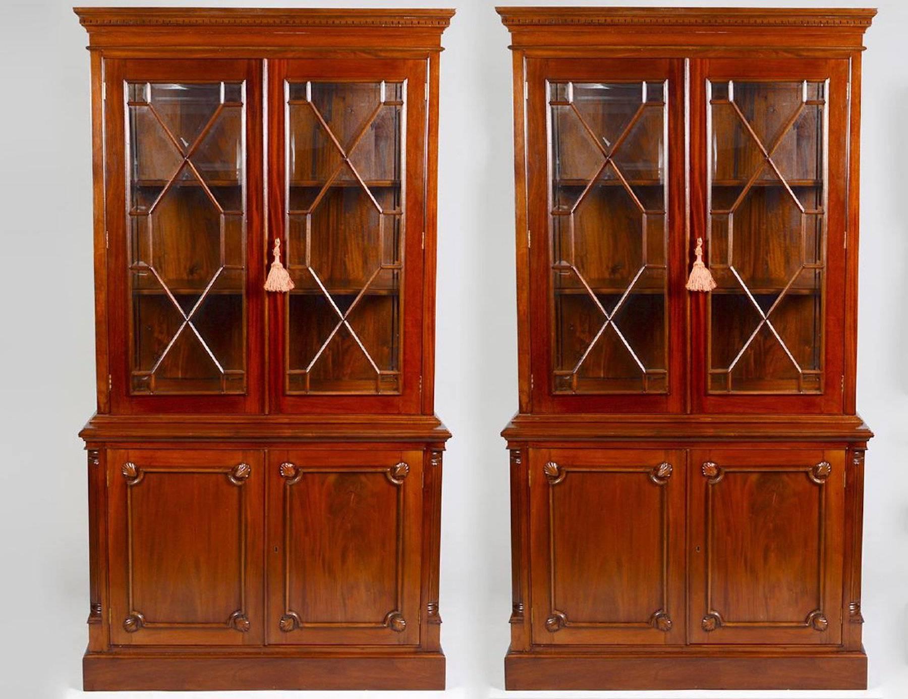Pair of English Georgian style mahogany bookcases or breakfronts, each one constructed in two pieces the upper cabinet with two mullioned doors and clear side panels, fitted with wood shelves. The lower case with a pair of shell carved paneled blind