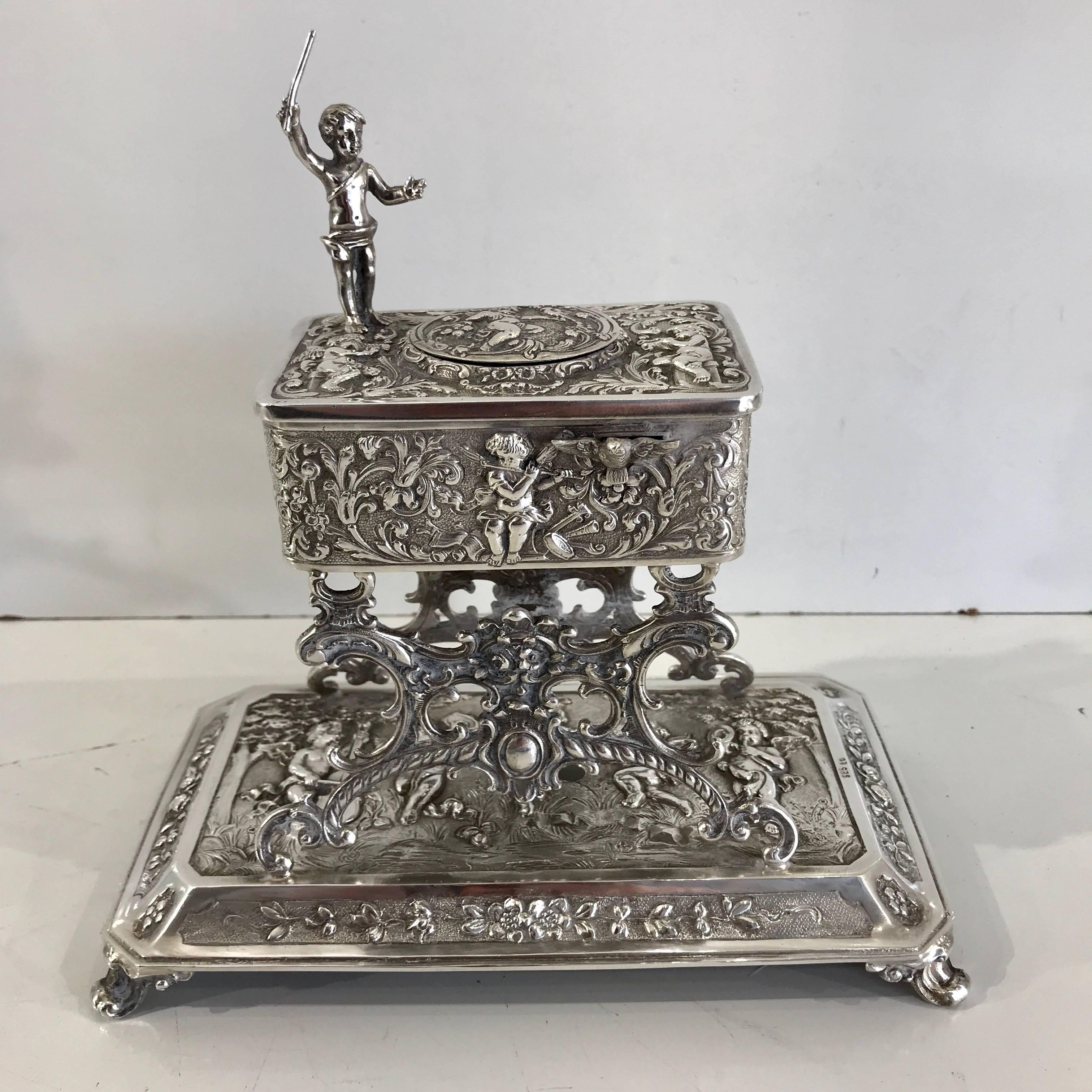 Large figural sterling singing bird table box. An elevated Rococo style rectangular bird box standing on four legs. The automaton bird is activated by a sliding lever with figural putto conductor above. When activated the vibrant miniature preening