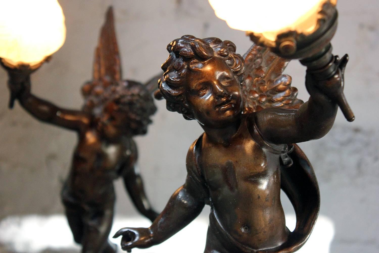 The well cast opposing pair of patinated spelter figural lamps, both with open wings, holding a flame glass shade, the other hand grasping a bow, one standing on a sphere with banding featuring zodiac signs with plaque reading 'L'amour Vagabond Par