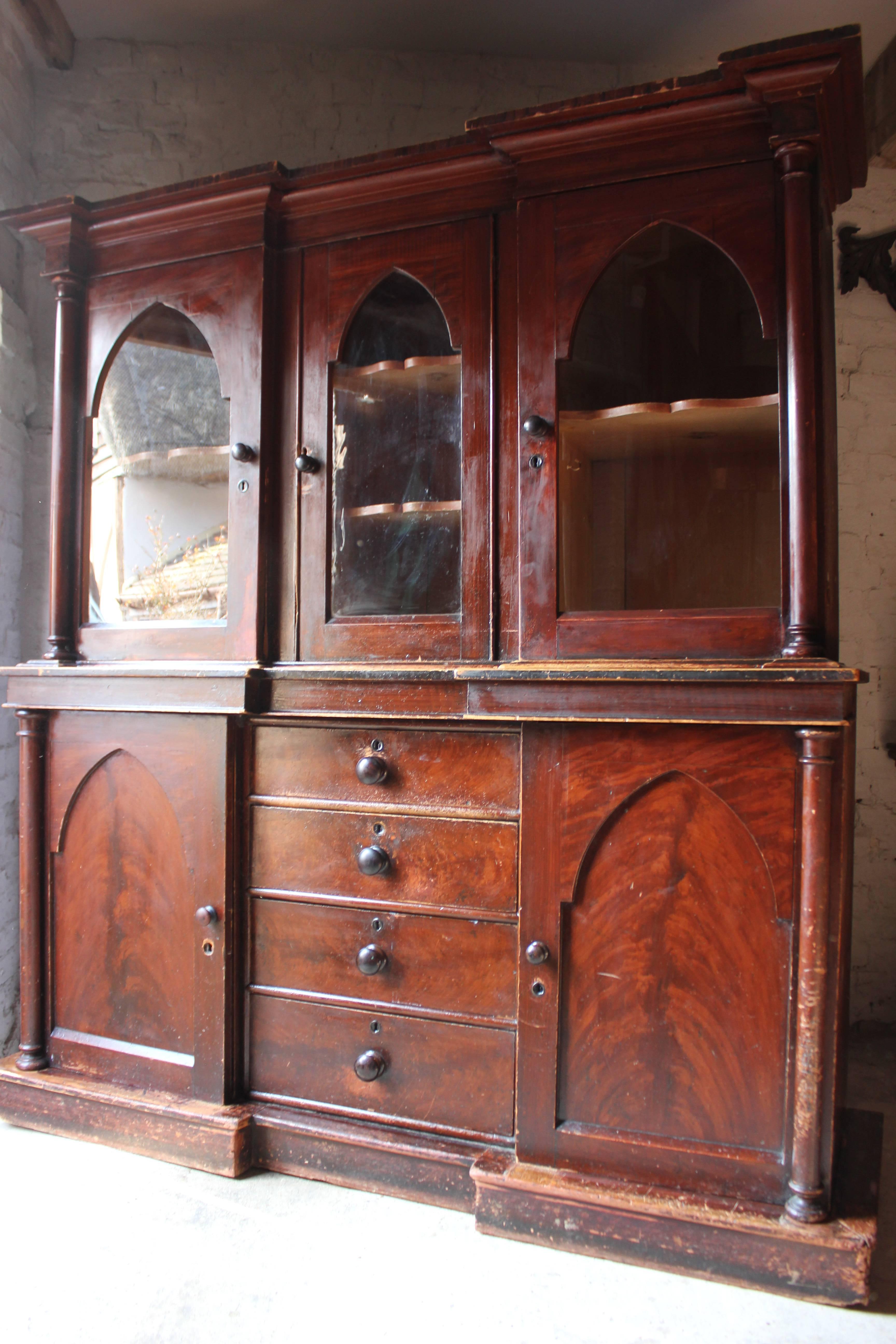 The handsome Provincial Gothic revival inverted breakfront bookcase, cupboard or cabinet, stained pine to simulate plum pudding mahogany, breaking down into three sections, the whole on a plinth base with pilasters to each flank and a stepped
