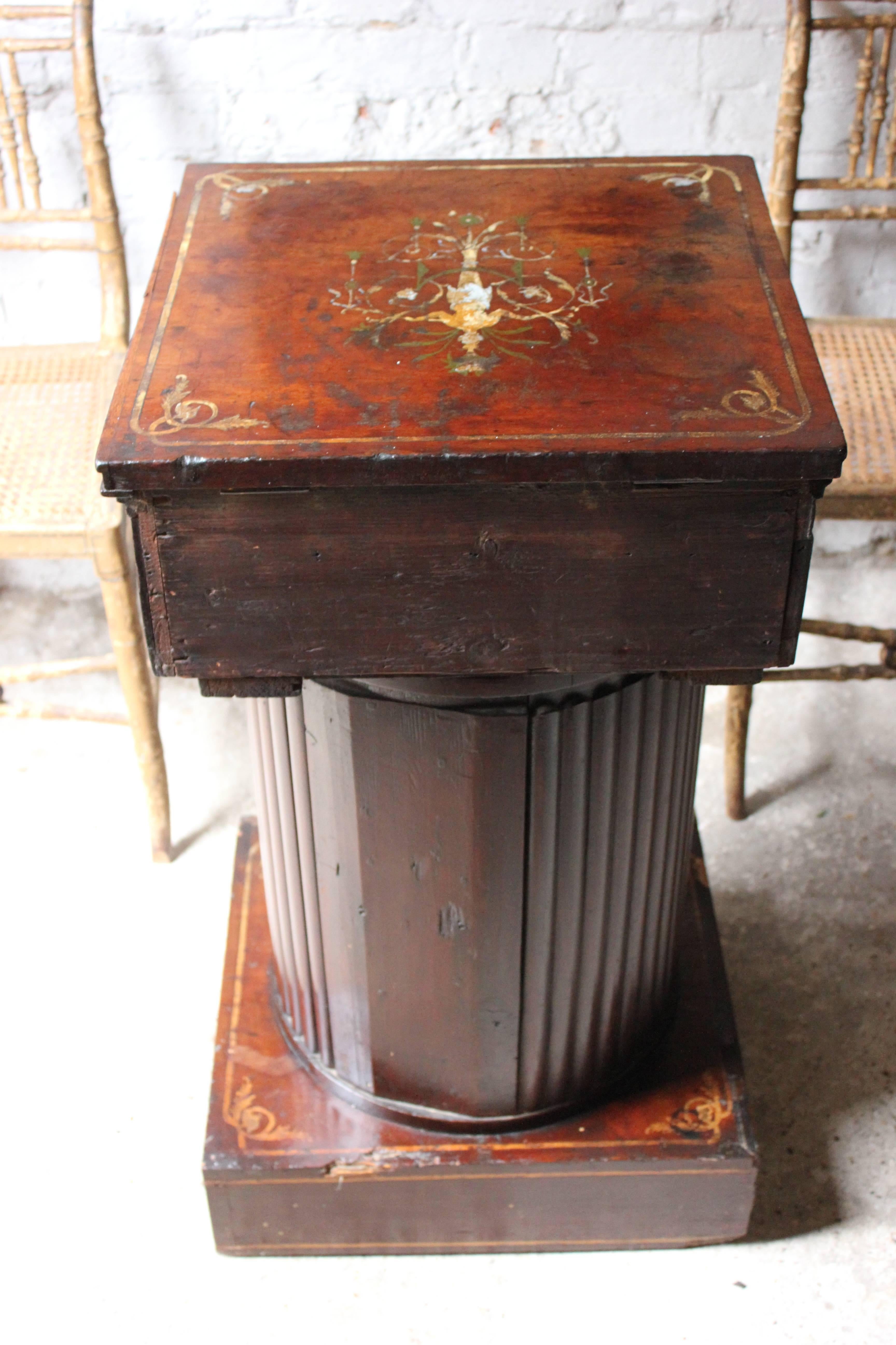 The painted mahogany pedestal cupboard of classical influence having a beautifully decorated square top showing a nymph amongst vines and swags, opening to reveal a vacant paper lined interior above a roundel carved and urn painted frieze, over a