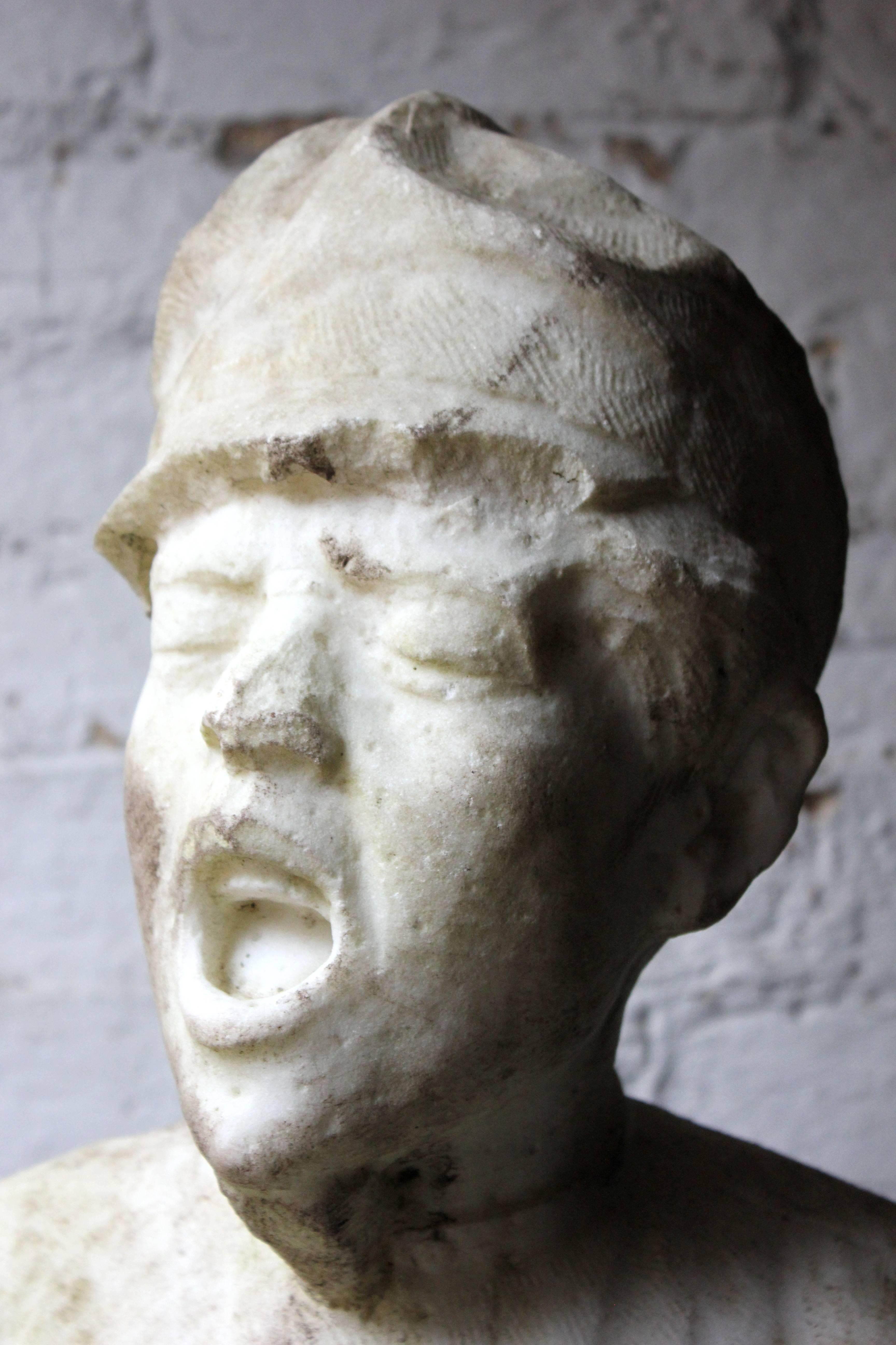 The evocative carved and weathered white marble bust showing the head and shoulders of a working class young boy, aged around ten, in a baker boy or flat cap, his eyes closed and his mouth wide open in exclamation, surviving from the middle period