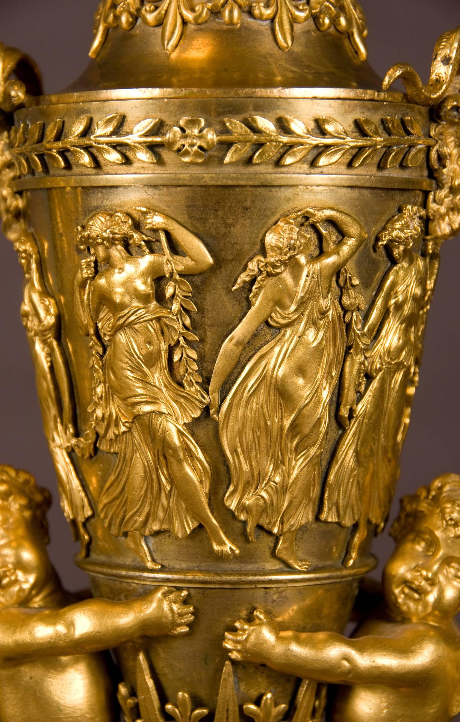 A rare pair of gilt bronze vases and covers each tapering ovoid urn with domed cover topped by a grape finial and flanked by rams' head masks, the body with scantly clad dancing maenads, supported by winged putti, on rectangular stepped plinth with