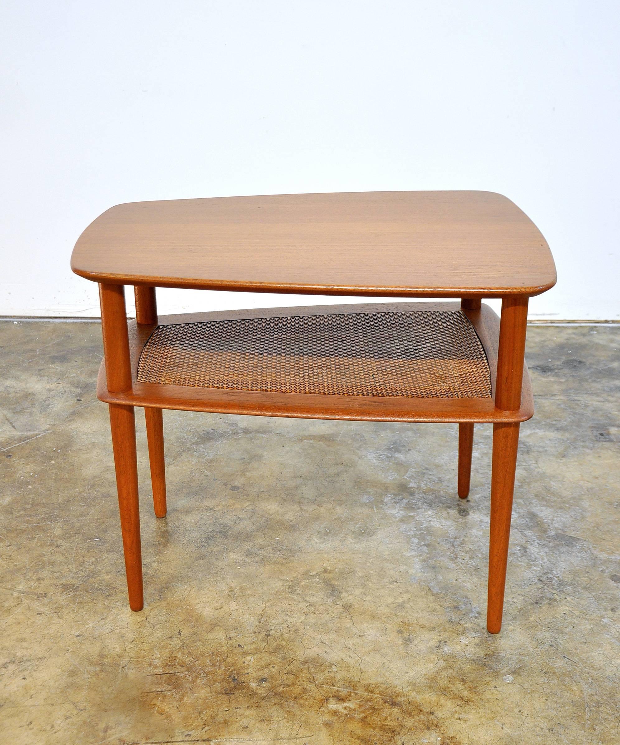Vintage Danish two-tiered solid teak end table with caned shelf. The table's classic Mid-Century design paired with its shape and size make it so versatile it can be used virtually anywhere. Beautifully grained teakwood. The top features a boat