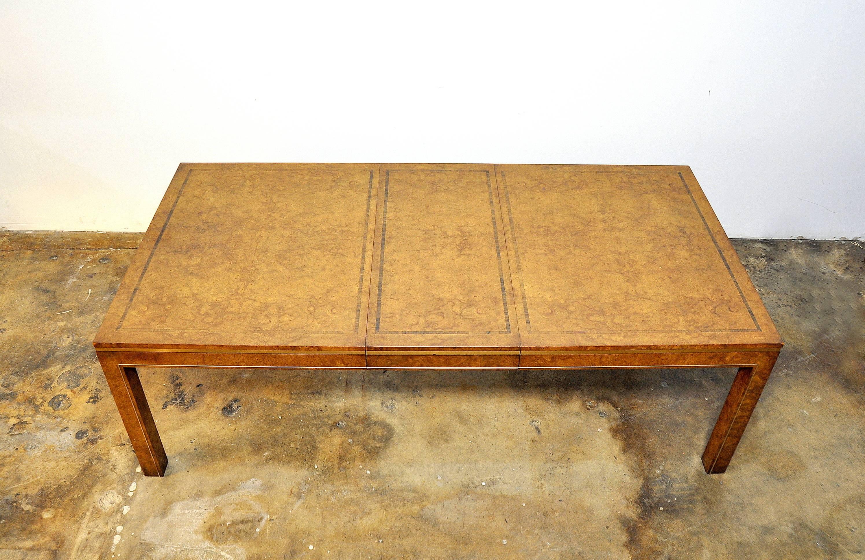 Elegant Mid-Century Modern expandable dining table featuring golden amboyna or maidou burl wood top with rosewood inlay. The table's skirt features brass banding, and the Parsons style legs are further accented by brass inlay. This Hollywood Regency