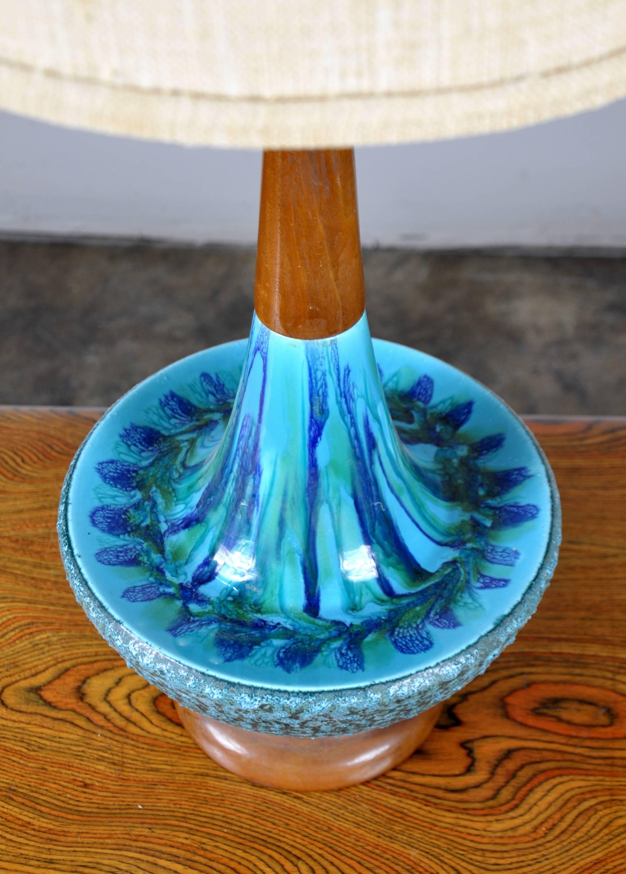 This tall Mid-Century vintage lamp dating from the 1960s features a smooth and textured ceramic body with drip glazed decoration in vivid turquoise and blue hues. The stem and base are wood. The tall original shade has an applied turquoise yarn