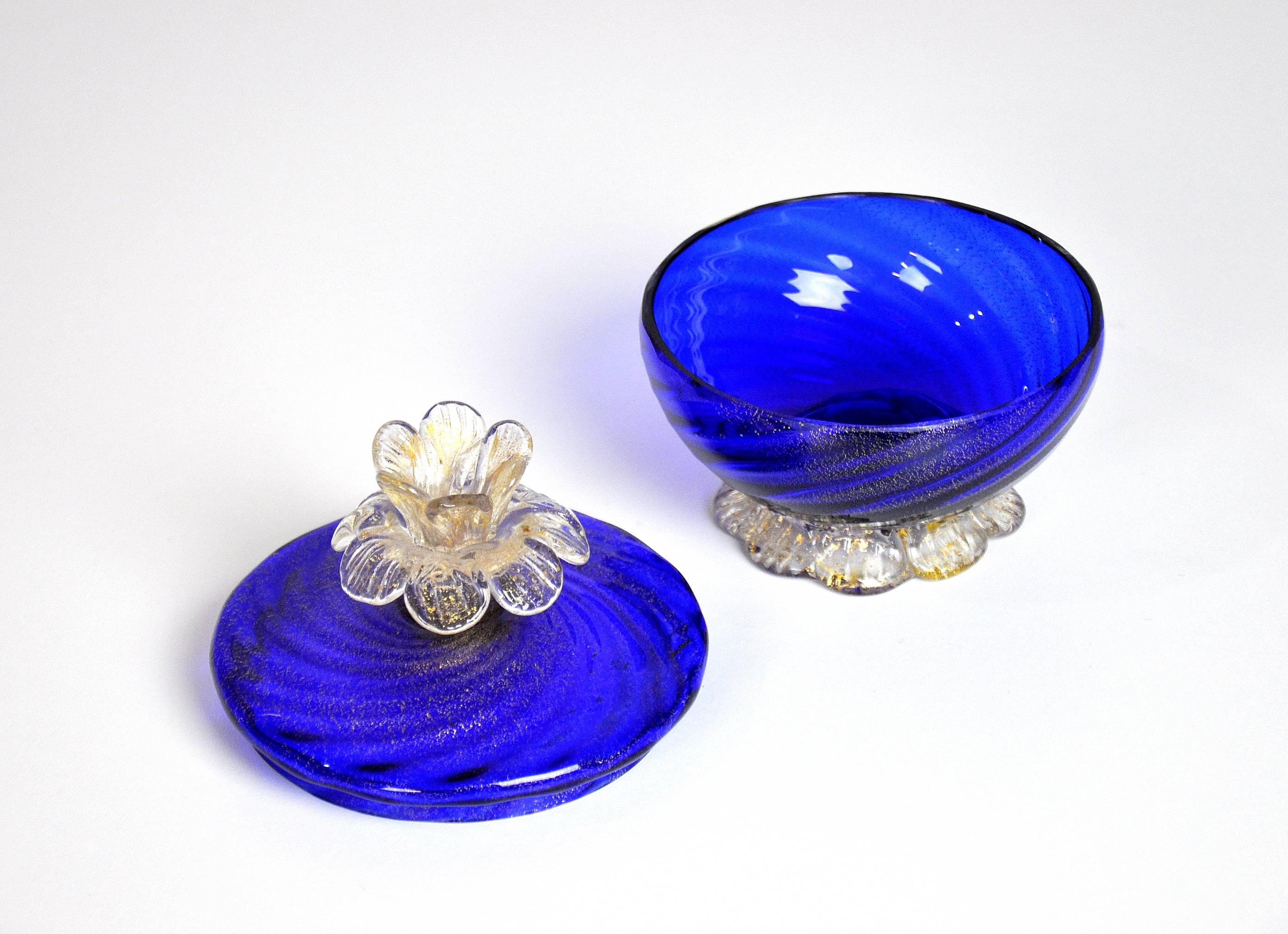 This vintage Venetian handblown art glass covered footed bowl features gold leaf inclusions, a figural finial skillfully shaped like a flower and a petal or leaf shaped base. The jar's deep, rich cobalt blue hue makes for a striking and elegant
