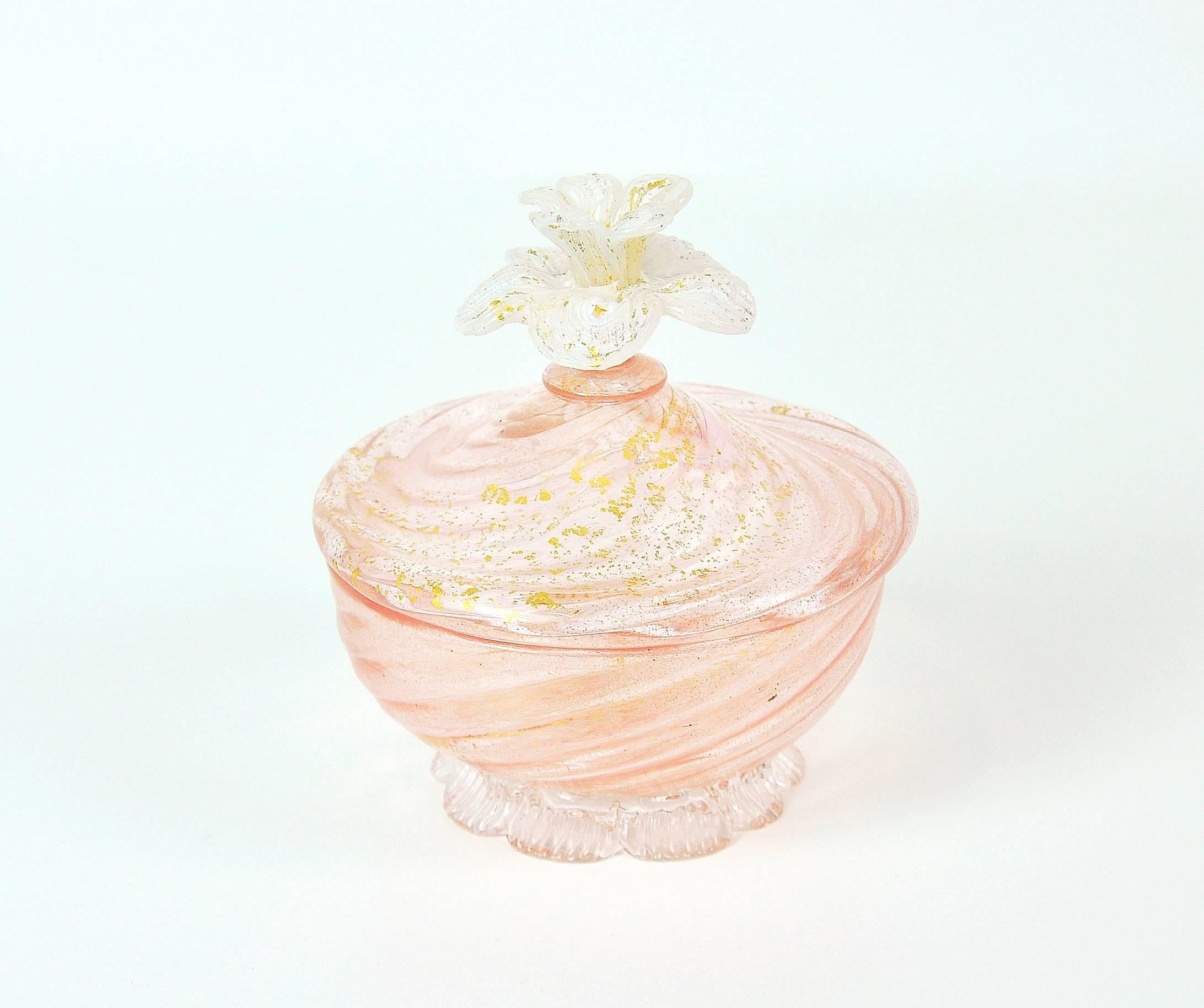 This vintage Venetian handblown art glass covered footed bowl features gold leaf inclusions, a figural finial skillfully shaped like a flower and a petal or leaf shaped base. The jar's soft, pastel pink hue makes for an elegant contrast to the gold