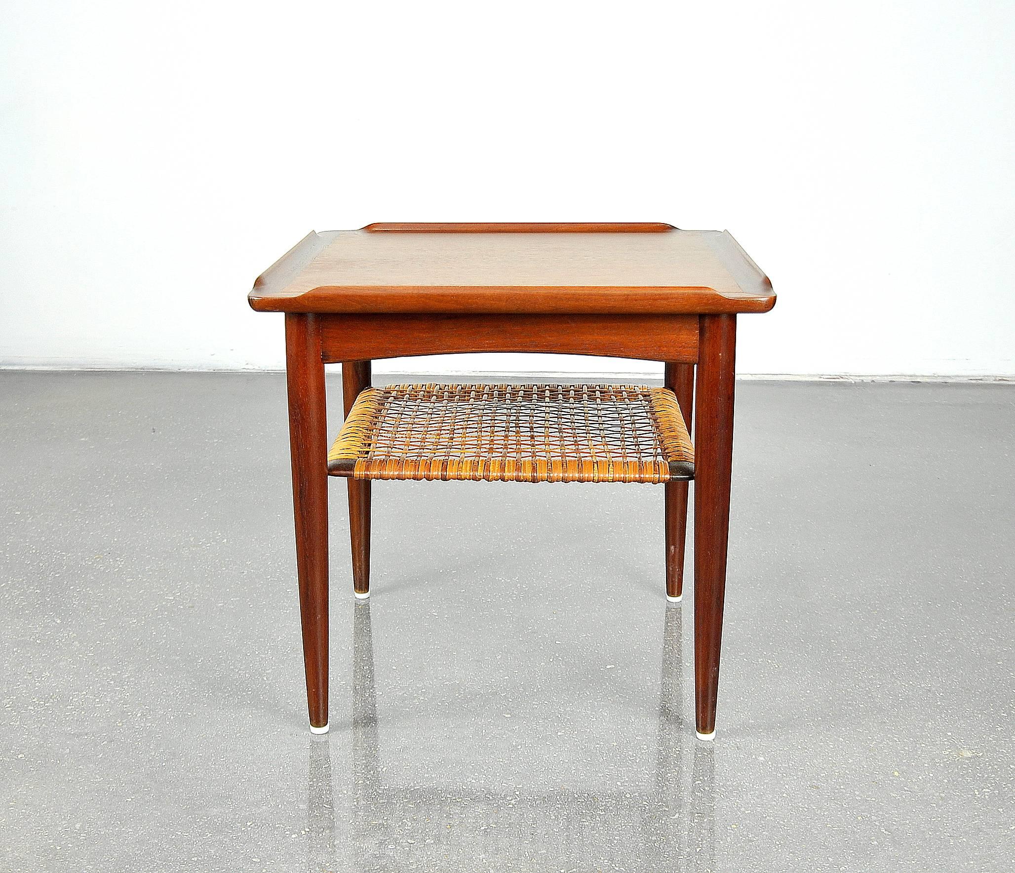 A pretty Danish modern square end table designed by Poul Jensen for Selig in the 1960s. This Mid-Century solid teak petite occasional table features sculpted raised edges and a caned magazine shelf that, being fitted with discreet brass hardware,