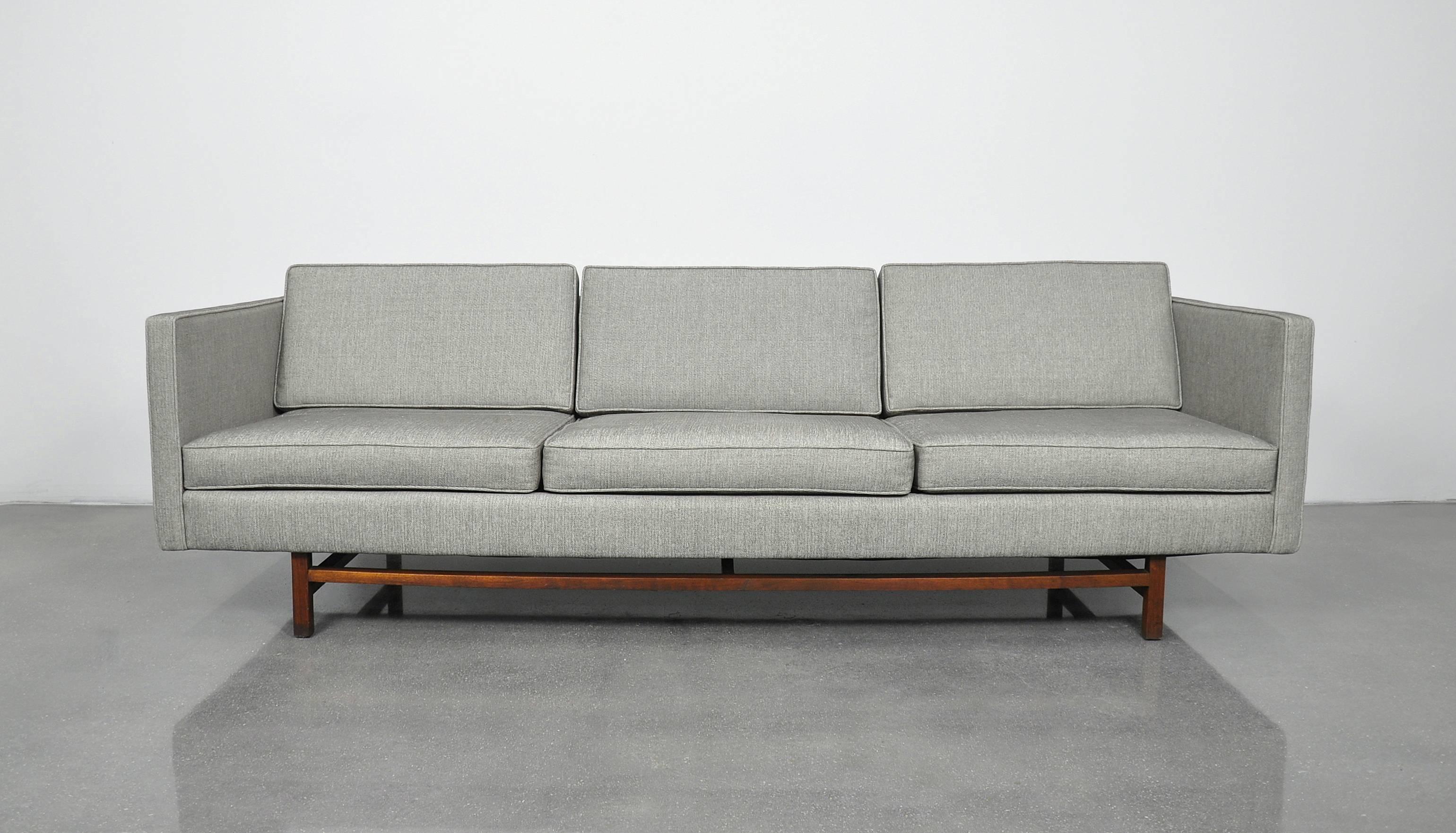 This reupholstered grey vintage sofa is a beautiful Mid-Century Modern example modeled in the style of Harvey Probber, and dating from the 1950s. The couch features a long and simple profile with walnut legs joined by stretchers. The sofa has
