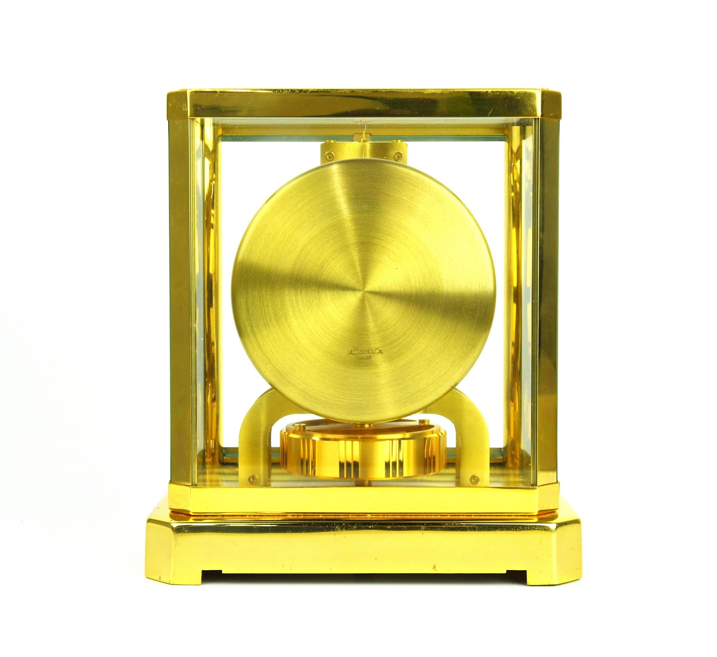 This perpetual motion Jaeger-LeCoultre mantle clock has a white dial, gold-toned Arabic numerals, hour markers and hands, and a gold-plated brass frame with clear glass windows. It does not need to be wound, as it gets the energy it needs to run