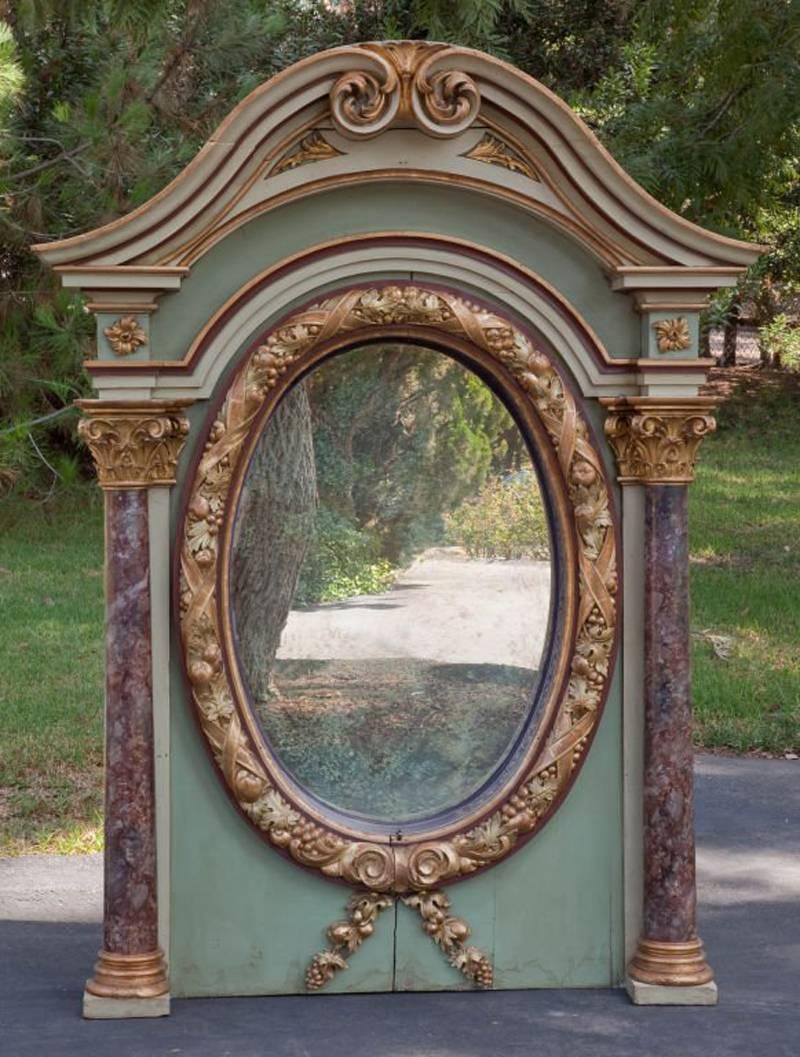 This superb early 19th century Italian architectural overmantel oval mirror is green painted with partial gilt. The curved pediment top is centered by double scrolls resting on Faux marble Corinthian columns flanking an oval plate bordered by a