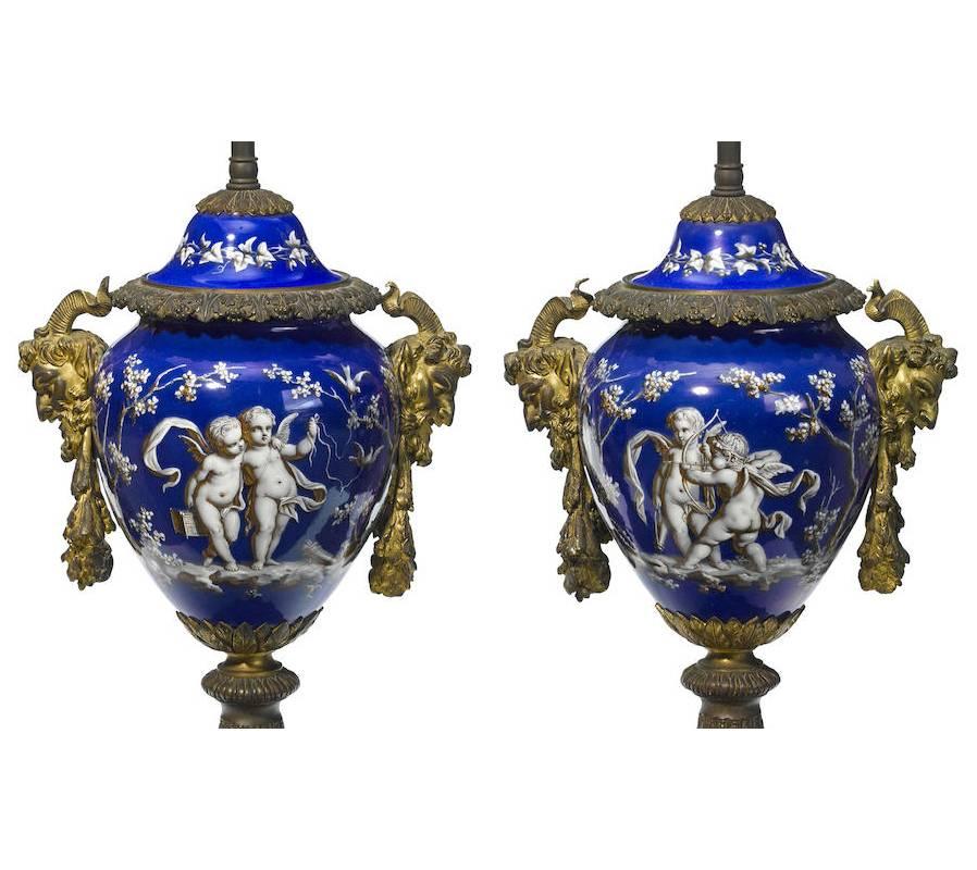 Beautiful 19th century pair of French gilt bronze and porcelain covered urns mounted to lamps. Rich blue porcelain urn portraying several playful cupid scenes flanked by ormolu masks and wreaths, all resting on gilt bronze base decorated with
