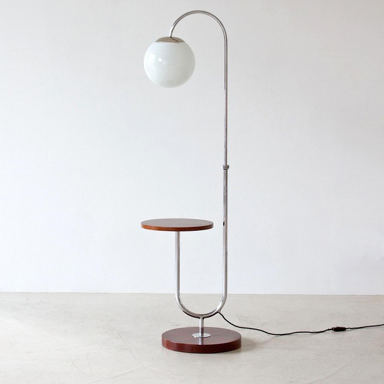 Lampadaire moderniste avec table basse intégrée. La structure tubulaire en acier est en métal nickelé, fixée sur un socle rond en bois et supportant une table basse arrondie sur un côté. L'ampoule en verre opalin peut être réglée en hauteur ou