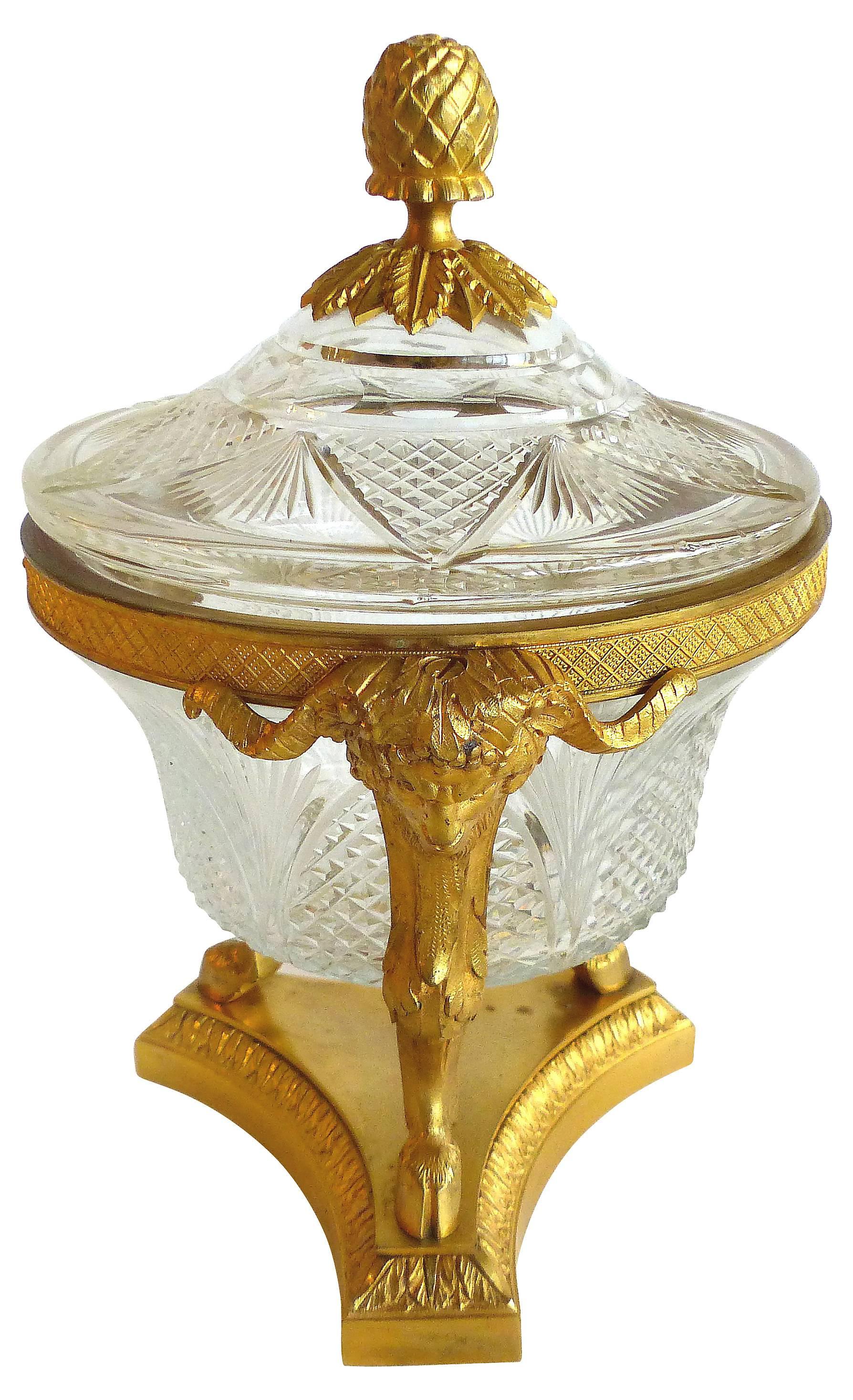 An elegant neoclassical Revival doré́ bronze lidded crystal bowl. Detailed with three fire-gilt ram heads joined by an ornate rim which encircles a cut crystal lidded bowl with a finely cast doré́ acorn finial. The legs rise from a classical tripod
