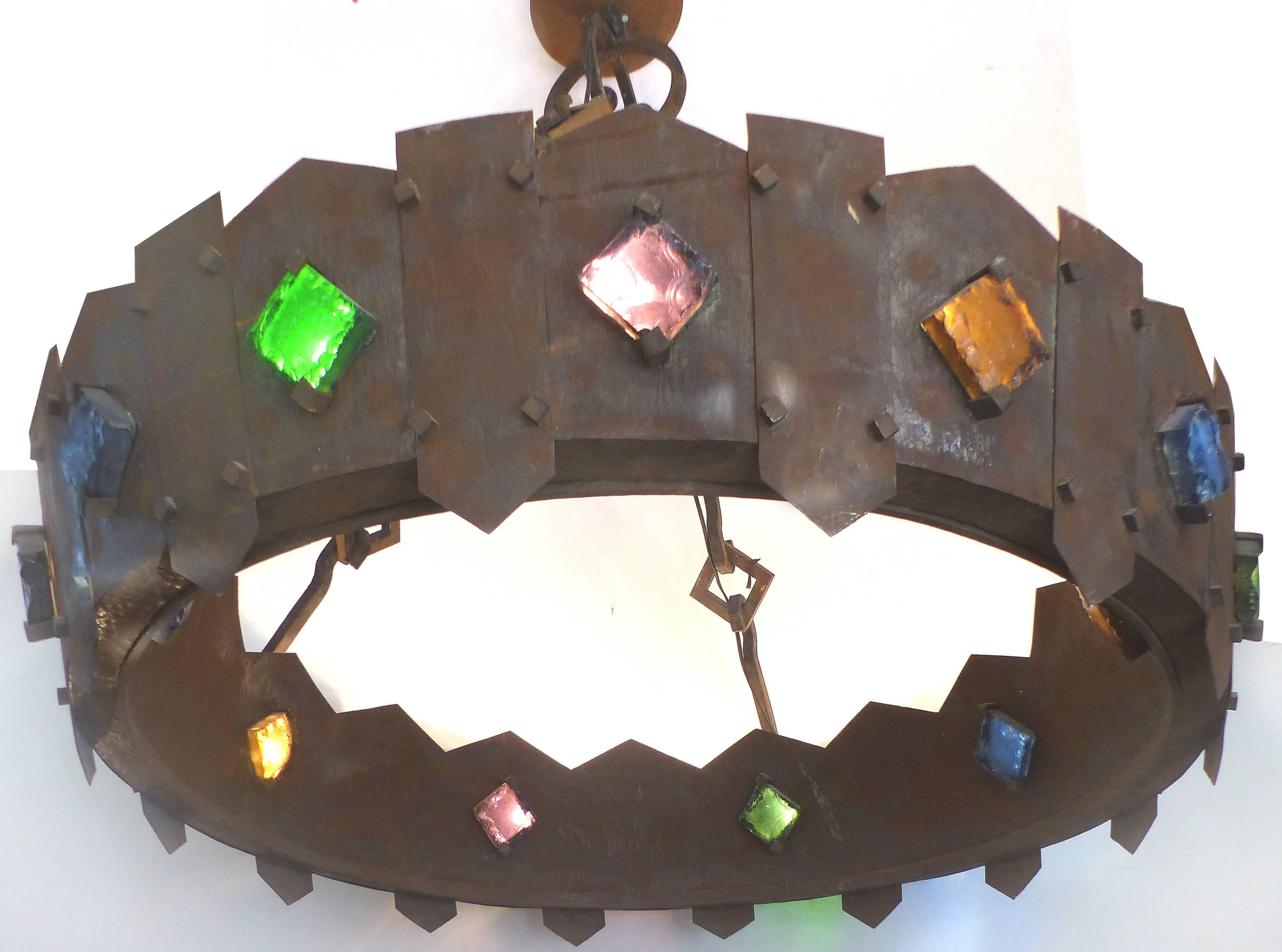 A monumental forged iron chandelier acquired in France created in the Gothic style. This wonderful fixture is wired with lights that illuminate upwards and through the colored slag glass mounted on the ring. The fixture is supported by a massive