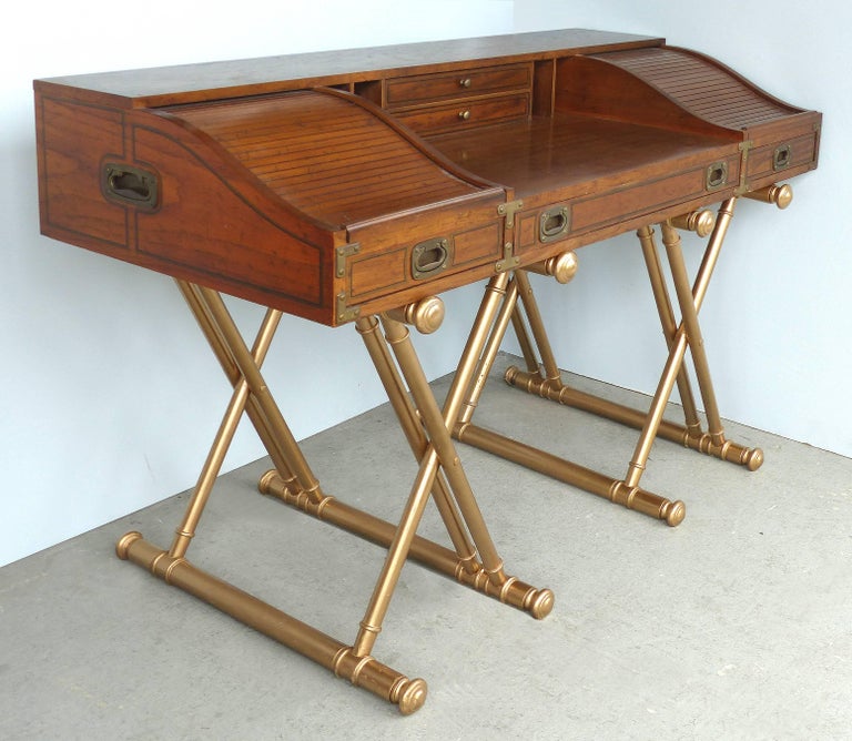 Offered for sale is a Campaign style desk designed by John Van Koert for Drexel. The desk is supported by gold crossed wood supports with a gilt finish. The top portion offers cubby-hole storage on either end which is hidden beneath rolling tambour